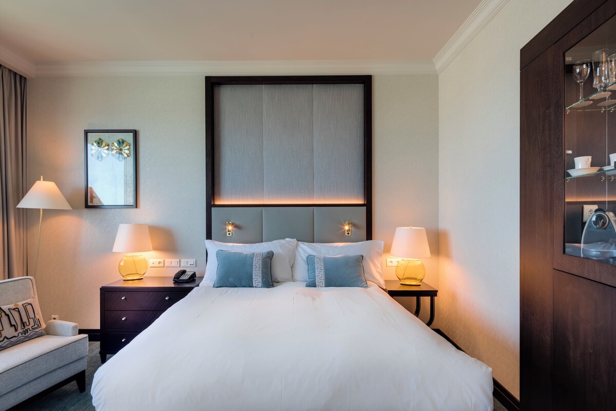 A neatly made bed with a headboard, two bedside tables with lamps, a small sofa with a cushion, and a wall-mounted cabinet with glassware.