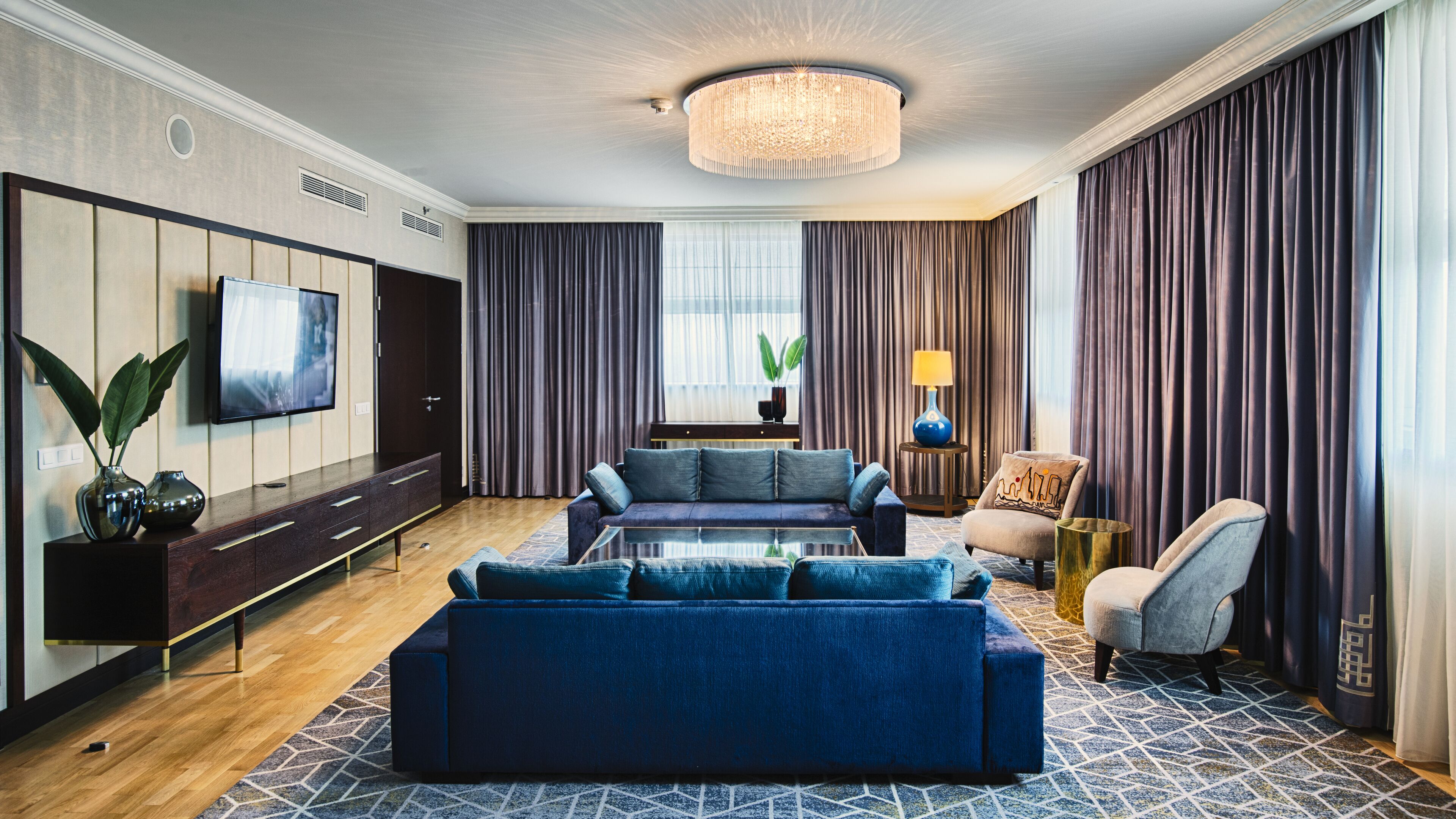 Ein modernes Wohnzimmer mit einer großen Couch, einem Couchtisch und einem an der Wand montierten Fernseher. Es gibt eine Chandelieber, eine Lampe und einen Topfpflanzen.