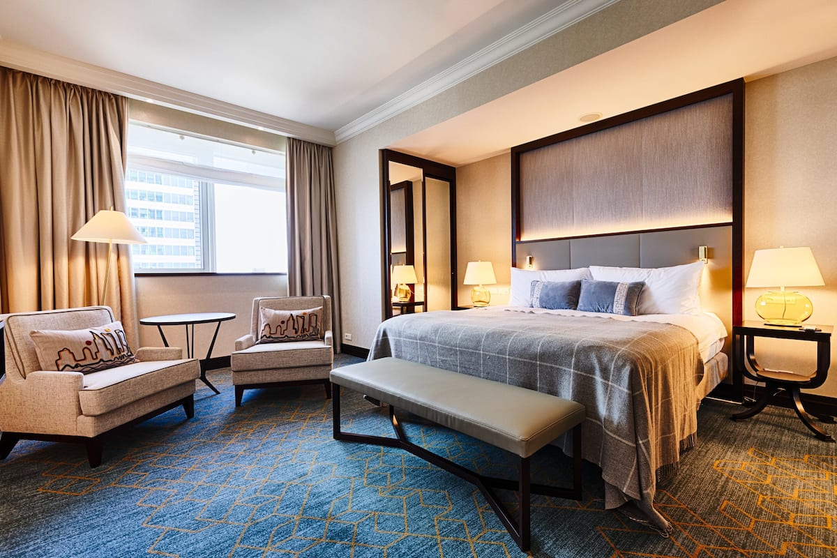 Una camera d'hotel con un letto grande, due poltrone, una panca, un tavolino e vista sulla città dalla finestra.