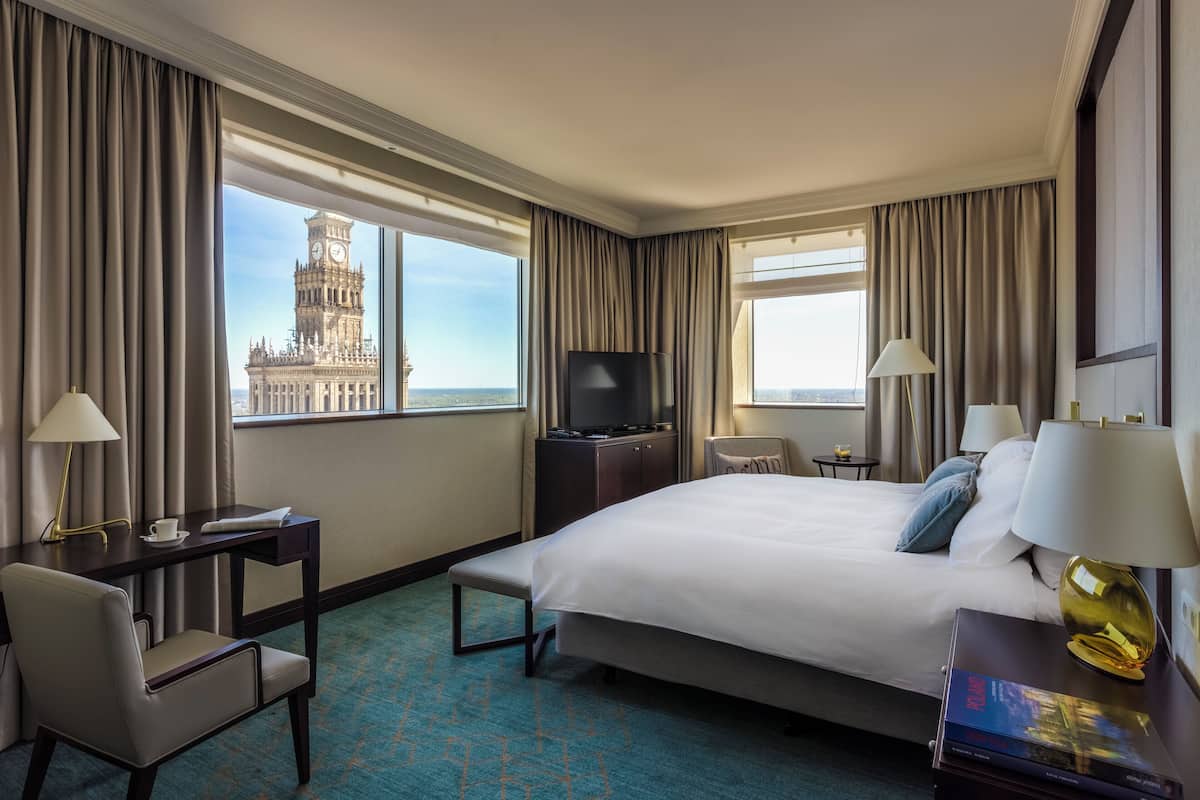 Una camera d'albergo con un letto grande, una scrivania con una lampada, una TV e vista su un campanile.
