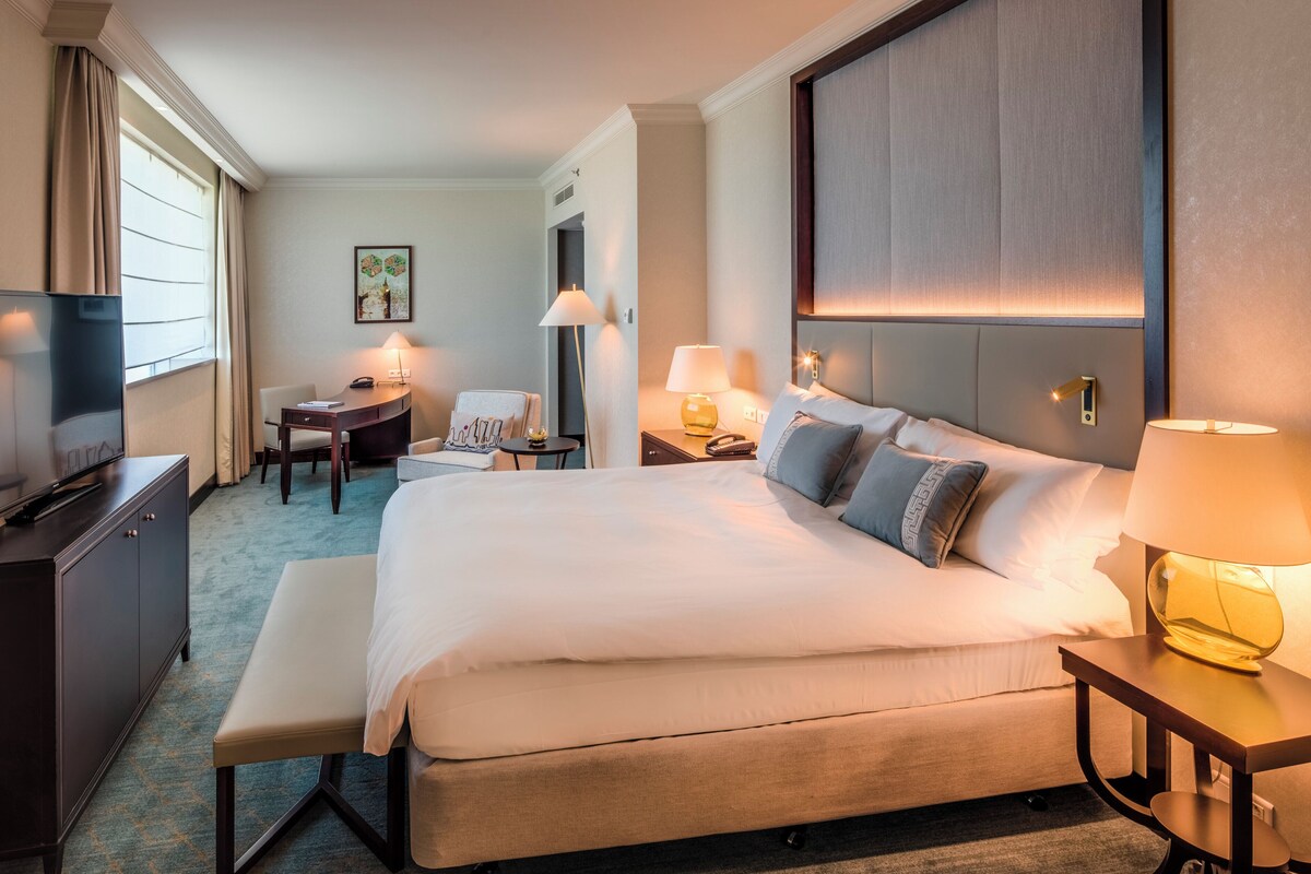 A hotel room with a large bed, bedside tables, a desk, a chair, and a television.