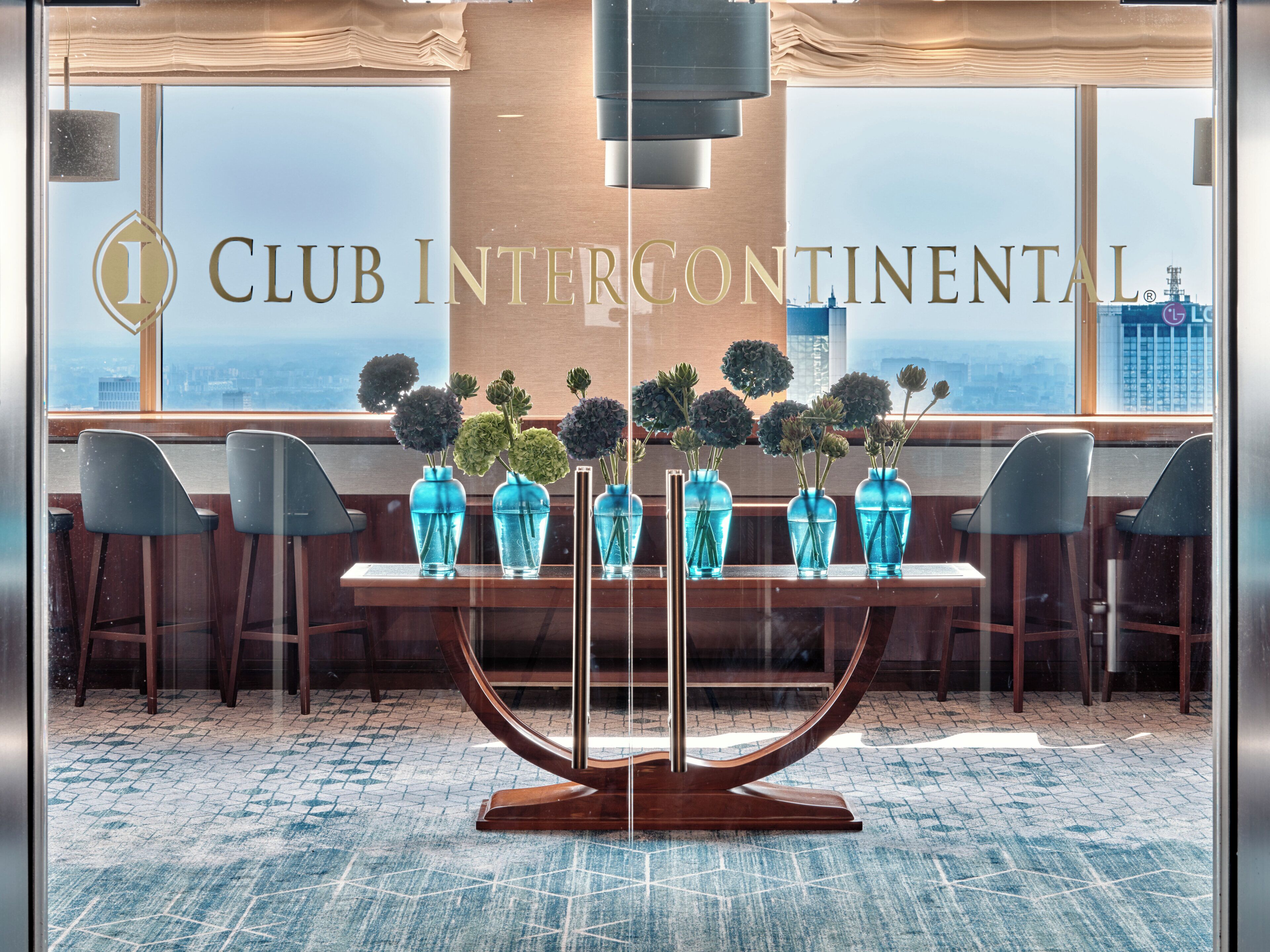 A modern interior with a table displaying blue vases with flowers, a large window with a view of the city, and the text "CLUB INTERCONTINENTAL" on the wall.