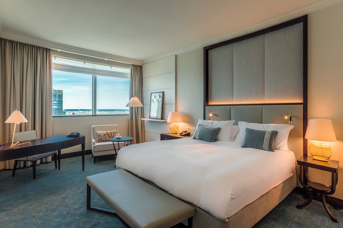 A hotel room with a large bed, a desk, a chair, a bench, and a view of the city through the window.