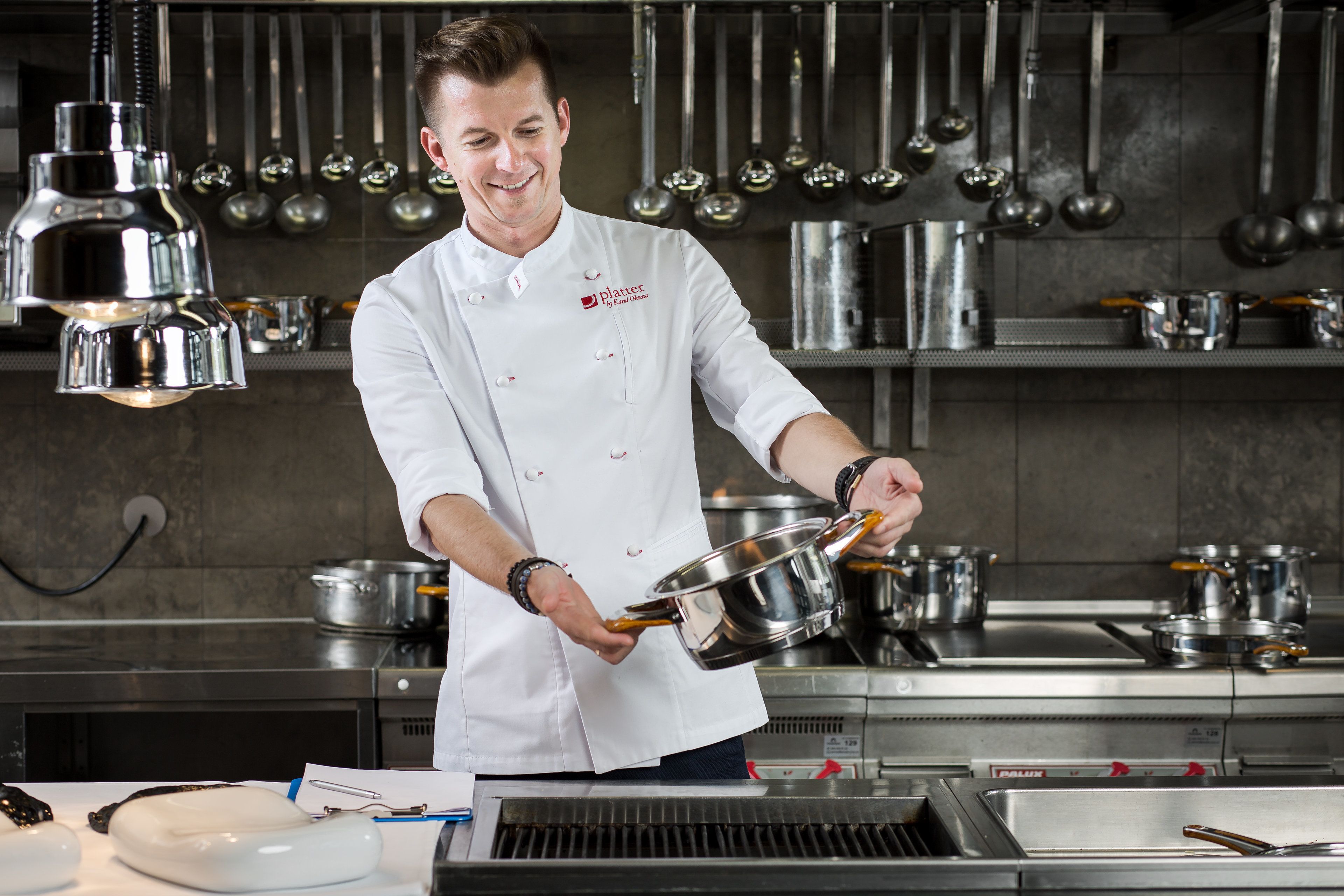 A chef in a professional kitchen with stainless steel equipment and hanging pots.