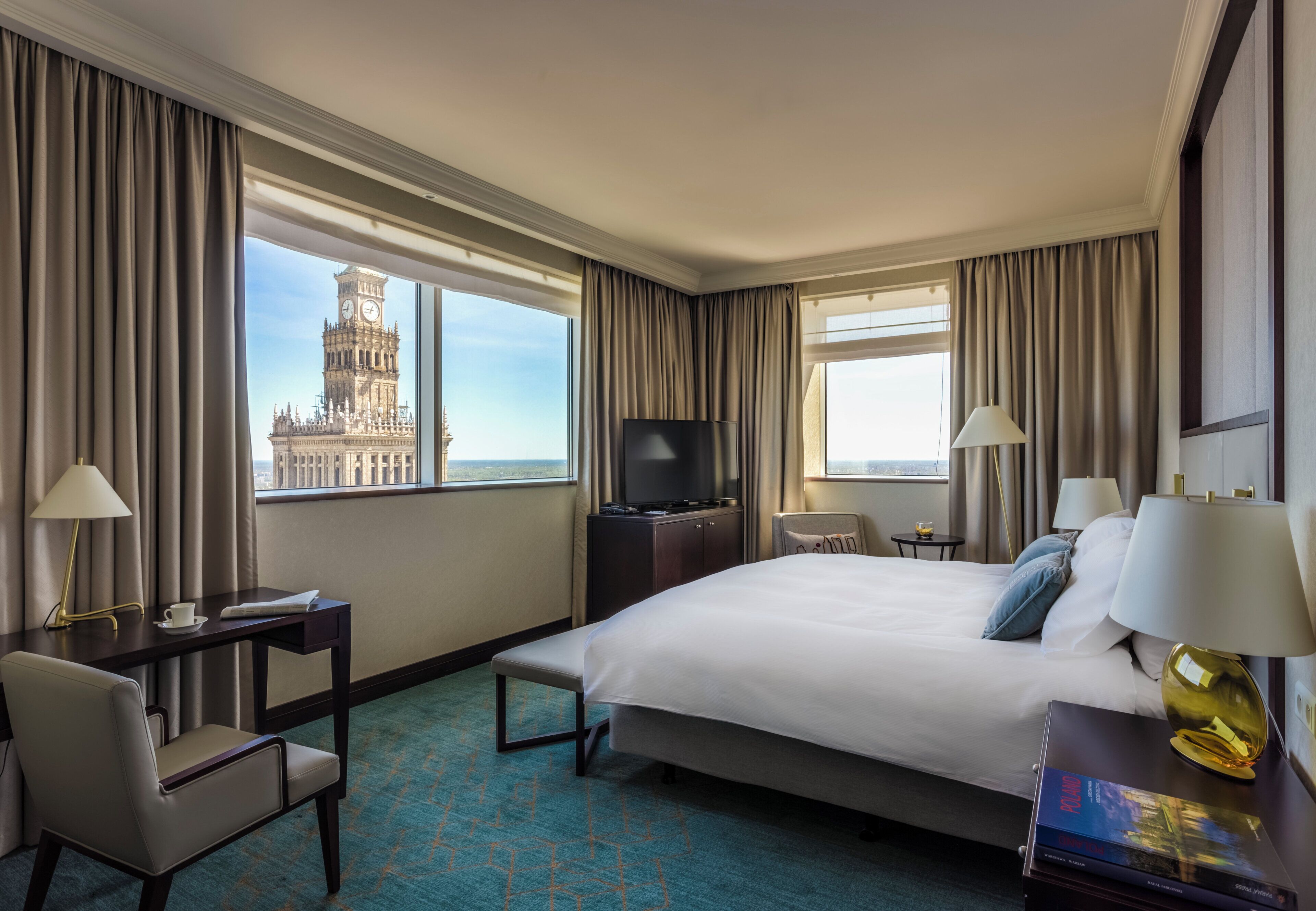 A hotel room with a large bed, a desk with a lamp, a chair, a TV, and a view of a historic tower.