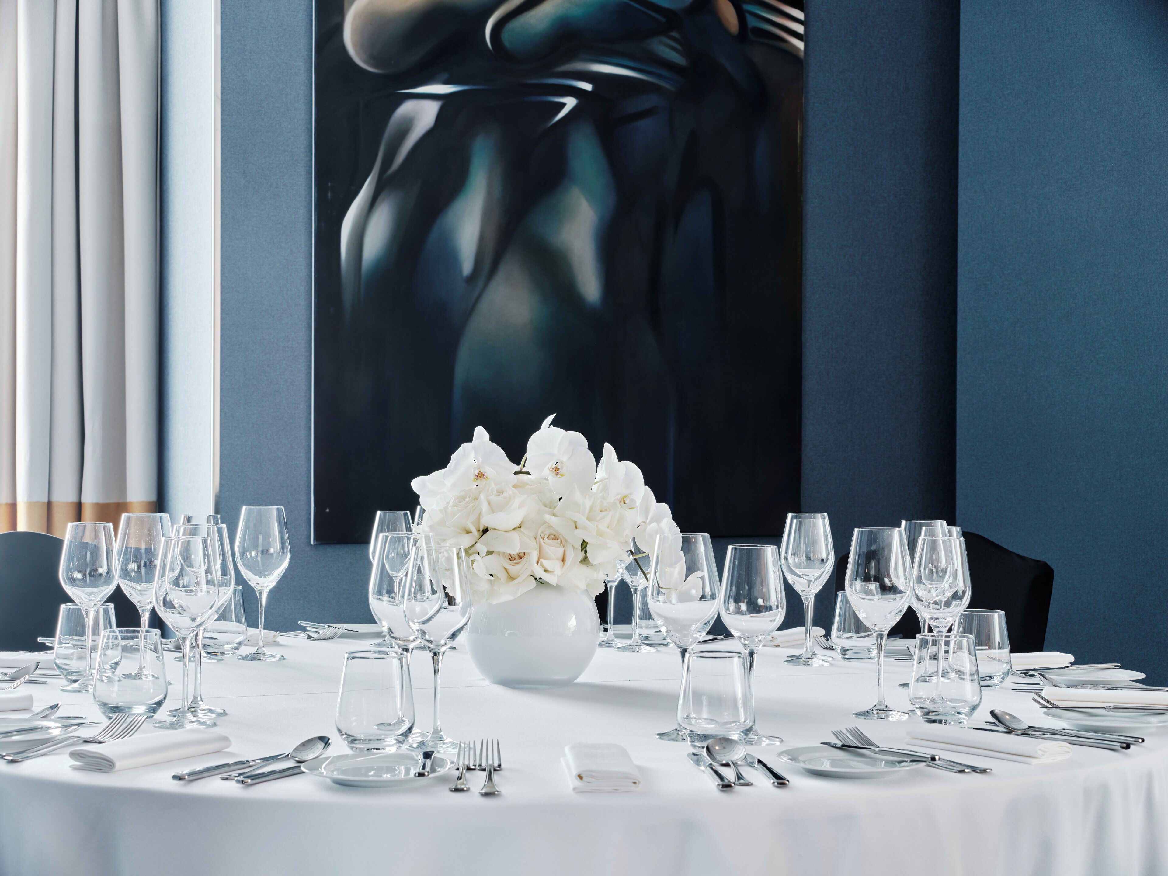 A dining table set with white tablecloth, silverware, and wine glasses, with a white floral centerpiece.