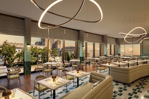 A spacious restaurant with multiple dining tables and chairs, large windows offering city views, and modern lighting fixtures.