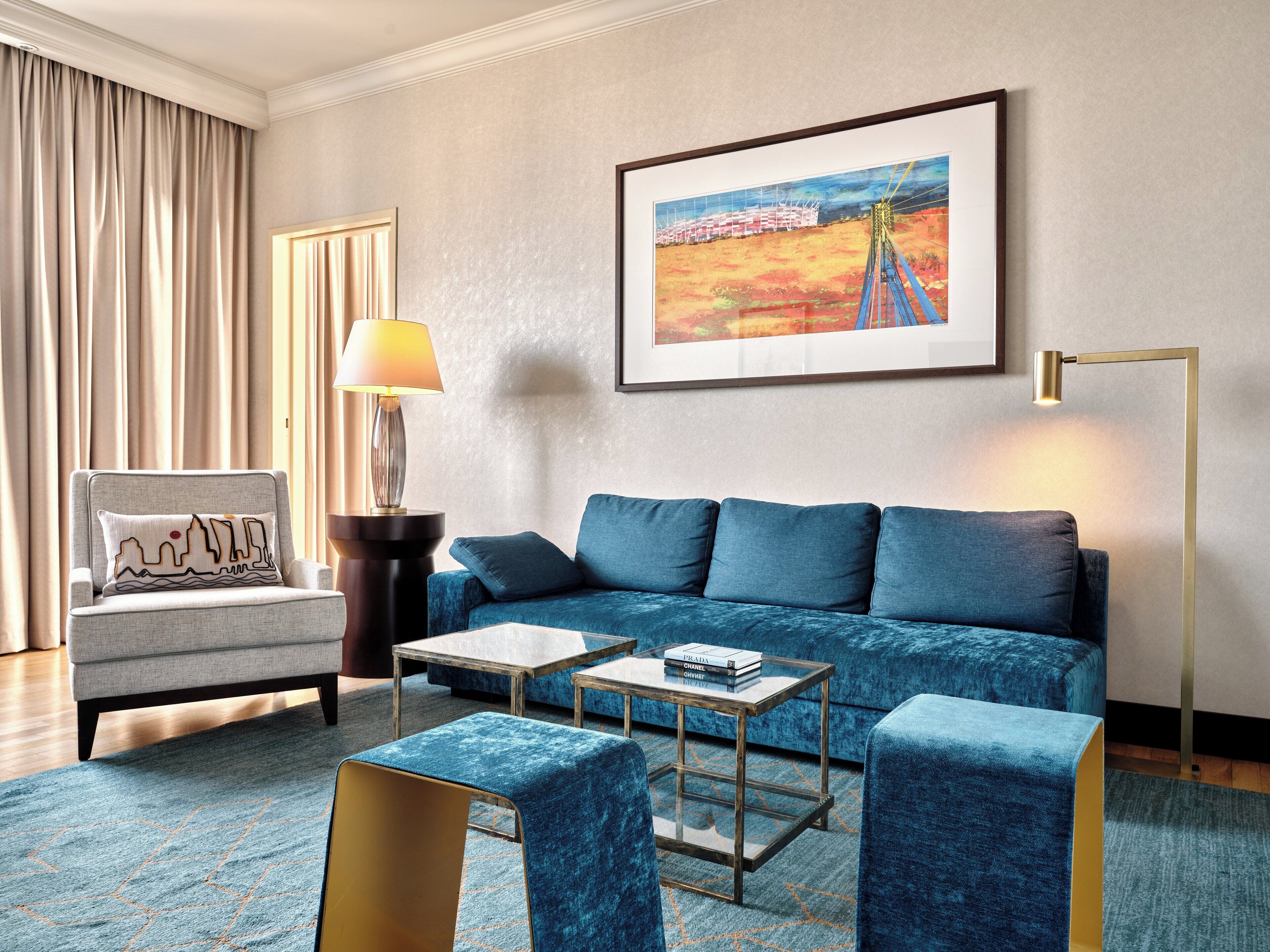 Ett modernt hotellrum med en blå soffa, en grå fåtölj, ett soffbord och en golvlampa.