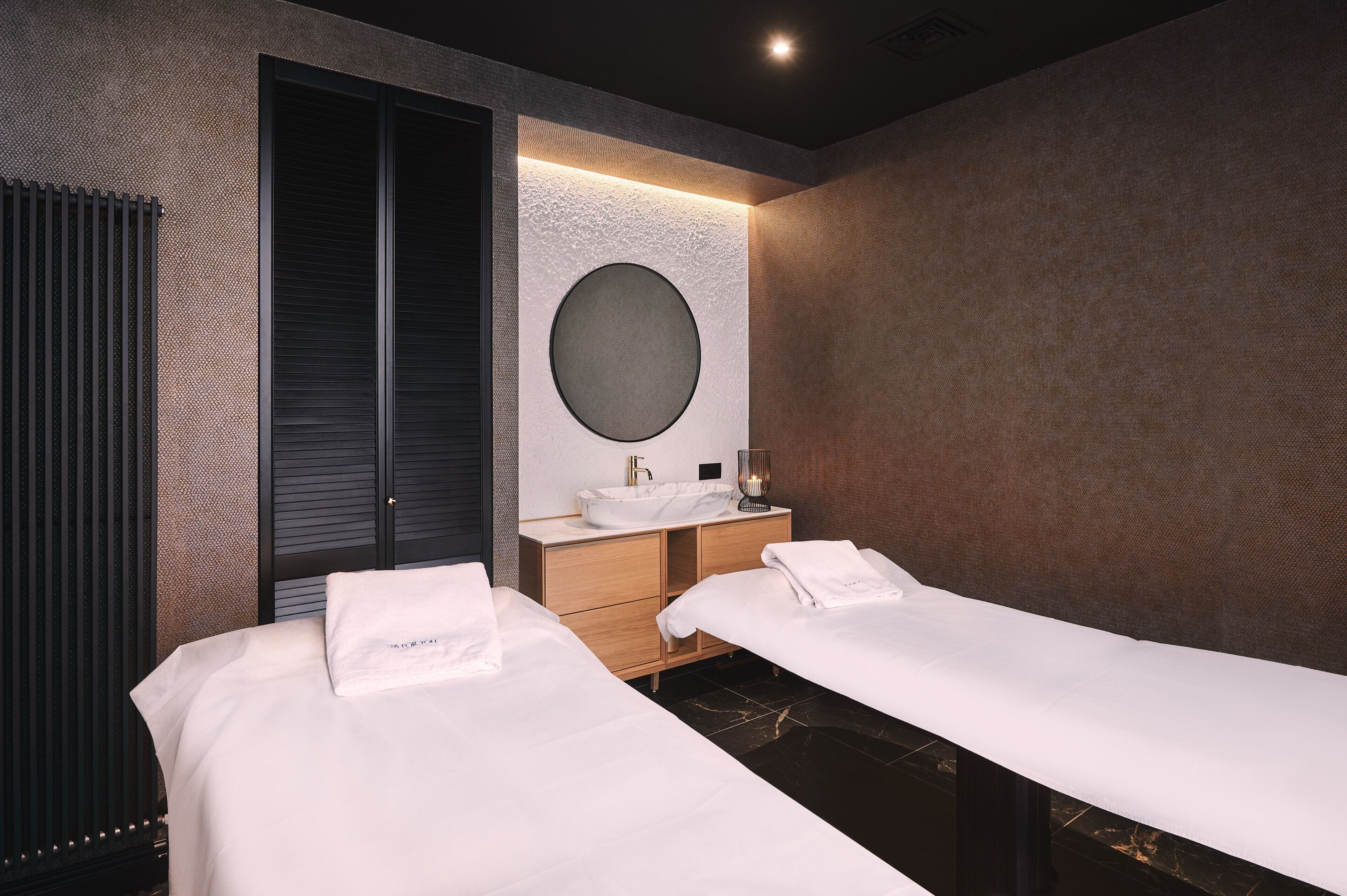 A modern hotel room with two single beds, a round mirror, a wooden vanity with a sink, and a dark-colored wardrobe.