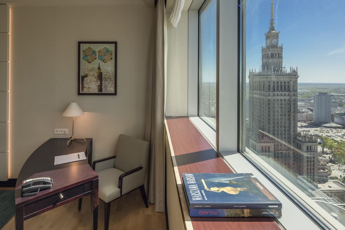 Camera d'albergo con scrivania, sedia e un'ampia finestra che offre vista su un paesaggio urbano e un edificio importante.