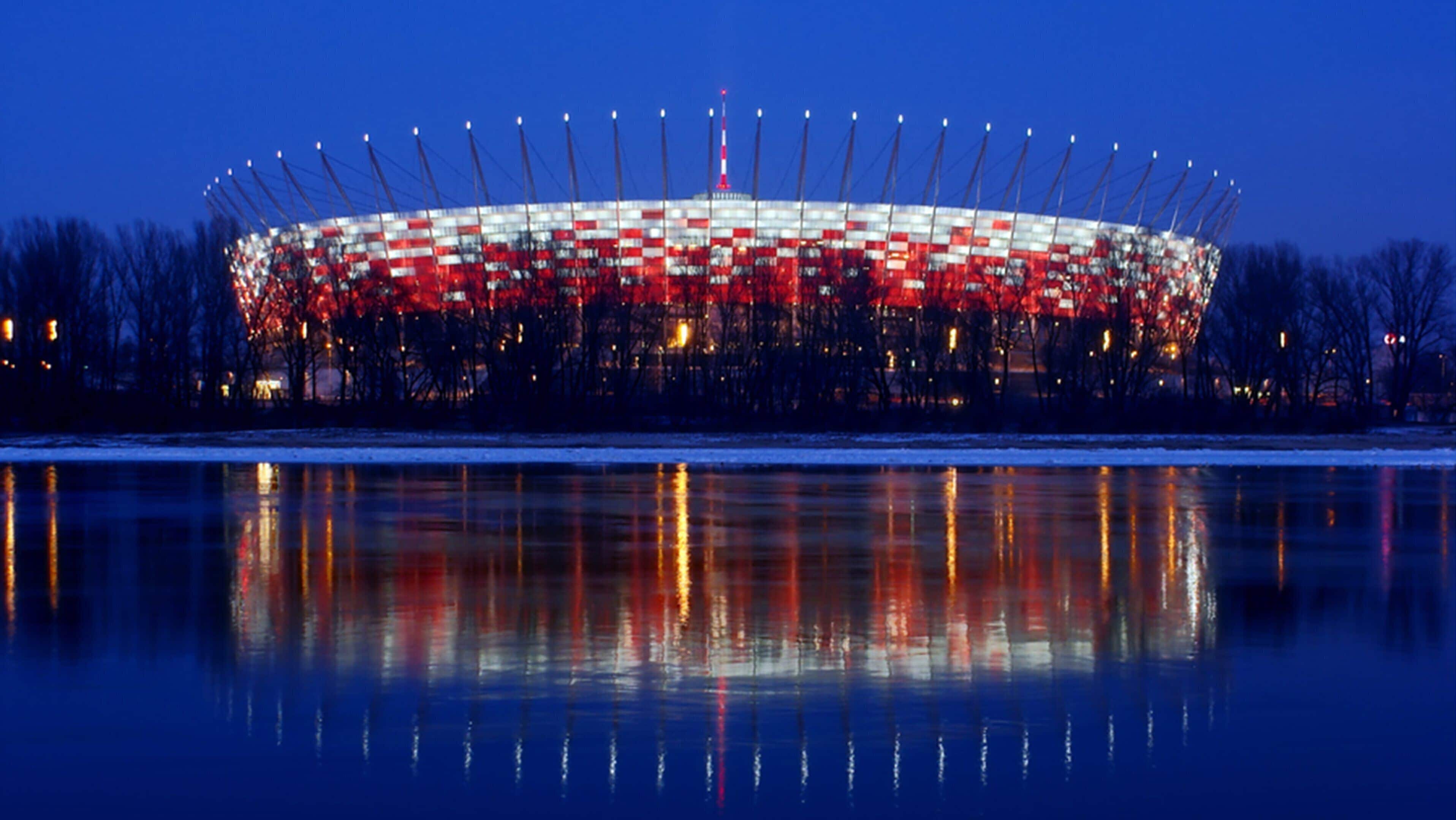 A large stadium with a circular roof and a central tower, surrounded by trees and a body of water reflecting its lights.