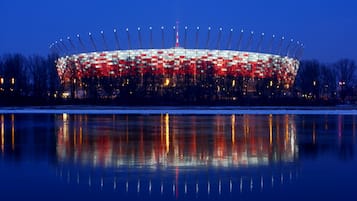 A large stadium with a circular roof and a central tower, surrounded by trees and a body of water reflecting its lights.