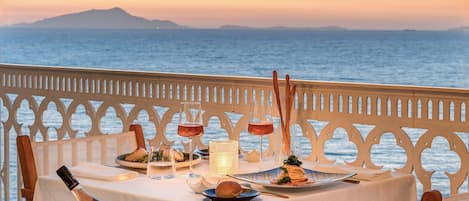 A table set for a meal with a bottle of wine, glasses, and a candle, overlooking a body of water at sunset.