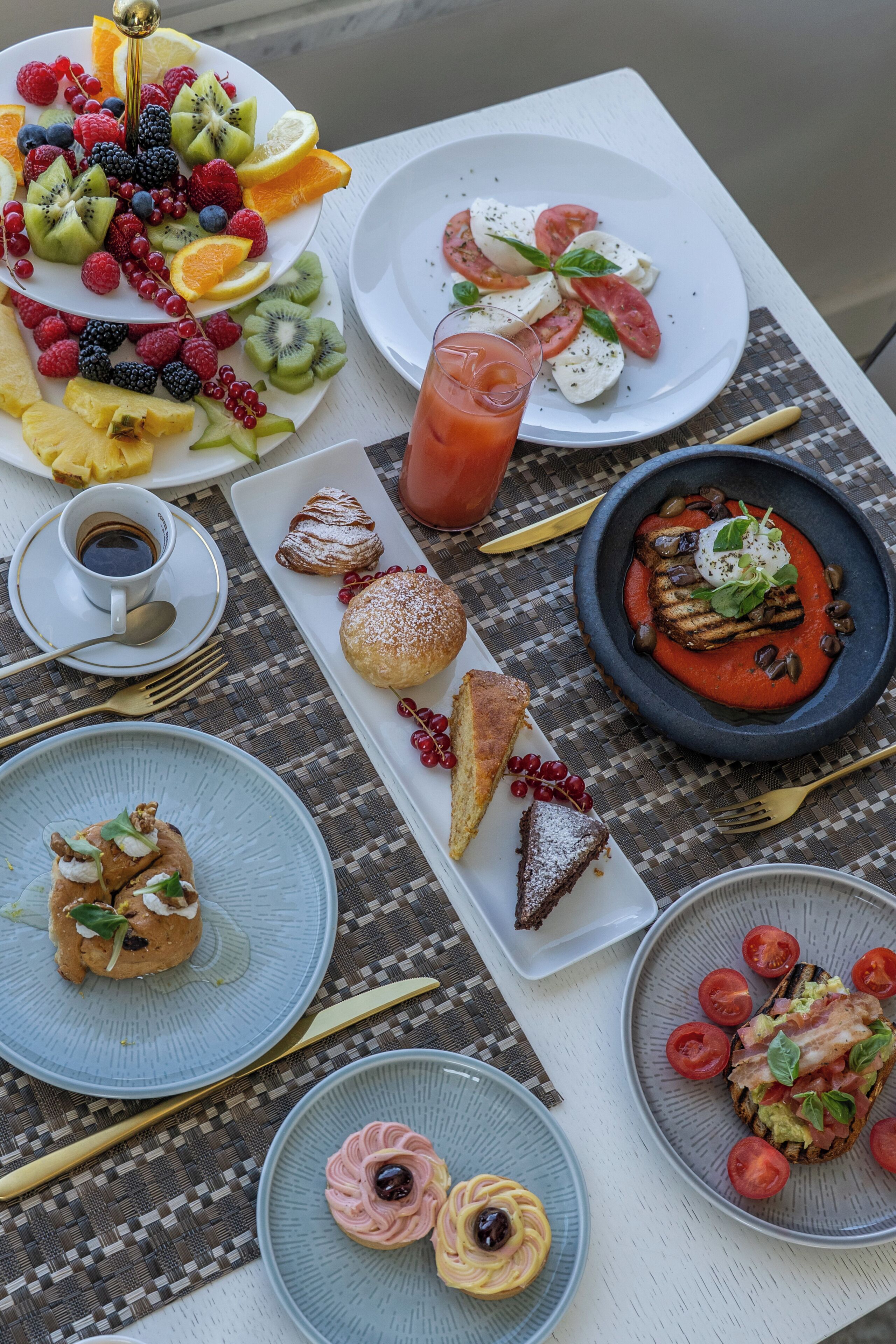 Ein Tisch reichhaltig gedeckt mit verschiedenen Speisen, darunter eine Obstplatte, ein Glas roter Saft, eine Tomate-Mozzarella-Anrichtung, ein Dessert mit Beeren und ein Teller mit roter Soße und Salat.