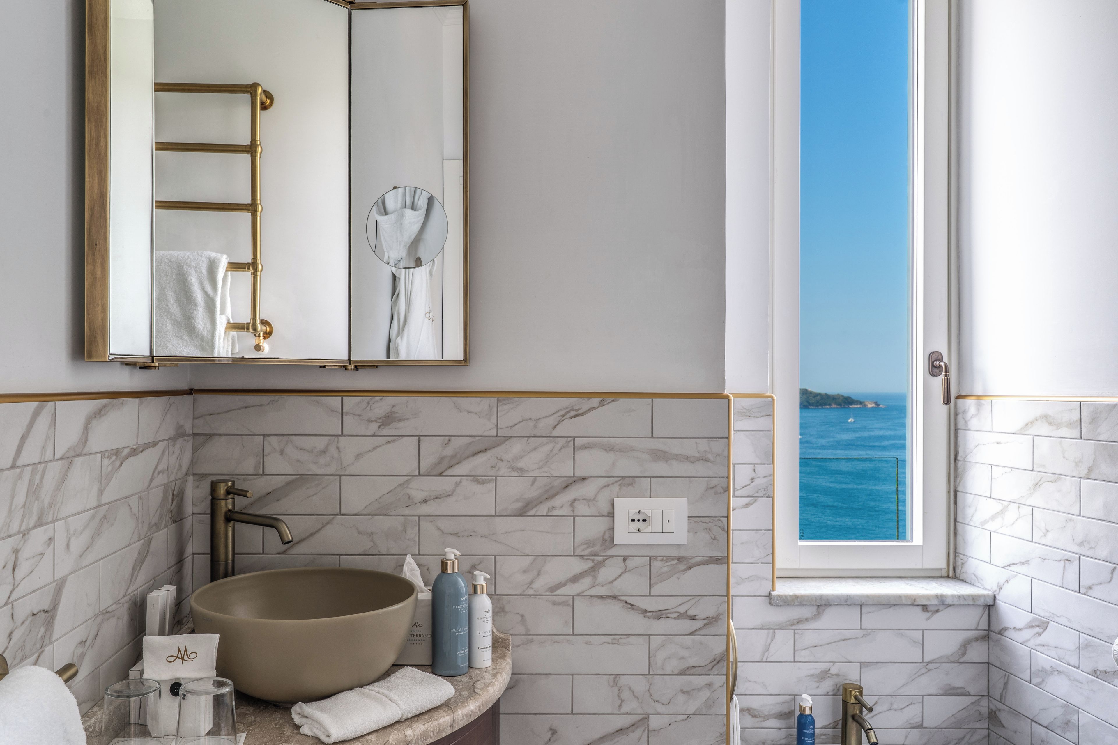 A bathroom with a large mirror, marble tiles, a sink with a bowl, and a window with a view of the sea.