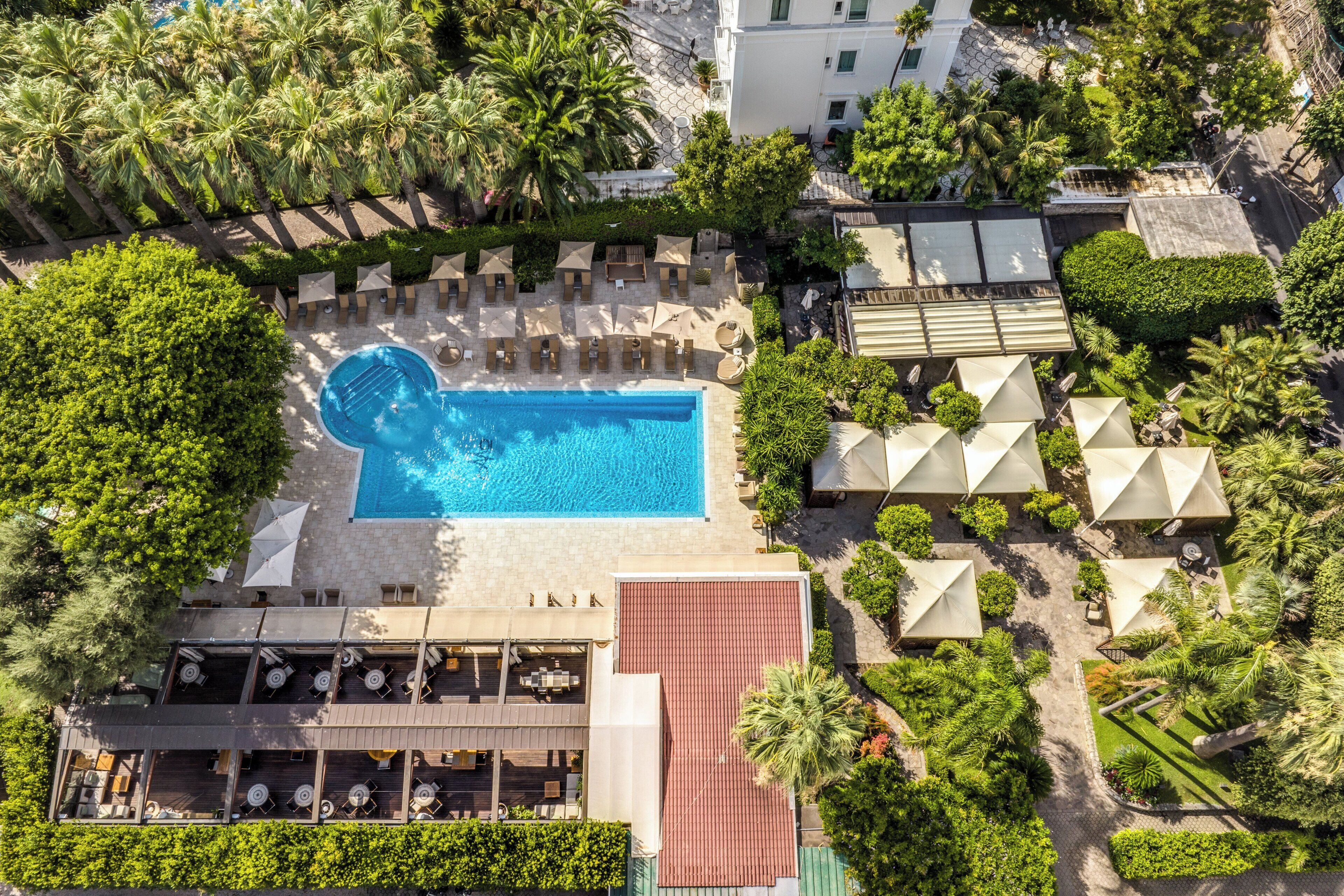 An aerial view of a resort with a swimming pool surrounded by lounge chairs and umbrellas, a covered dining area, and lush greenery.