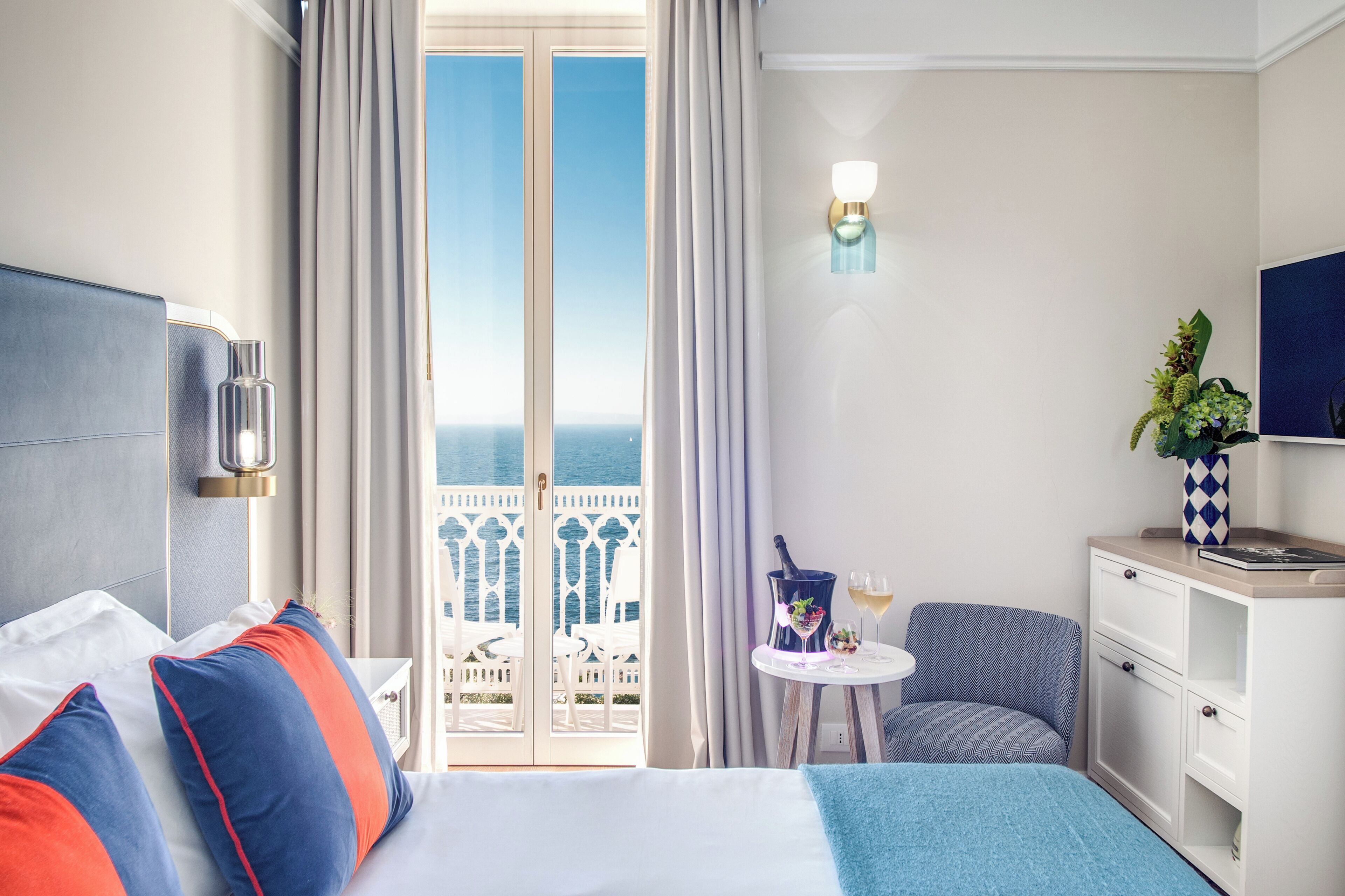 Habitación de hotel con cama, una silla, una mesita con una botella y vasos, una mesita de noche y vista al mar a través de una puerta de balcón.
