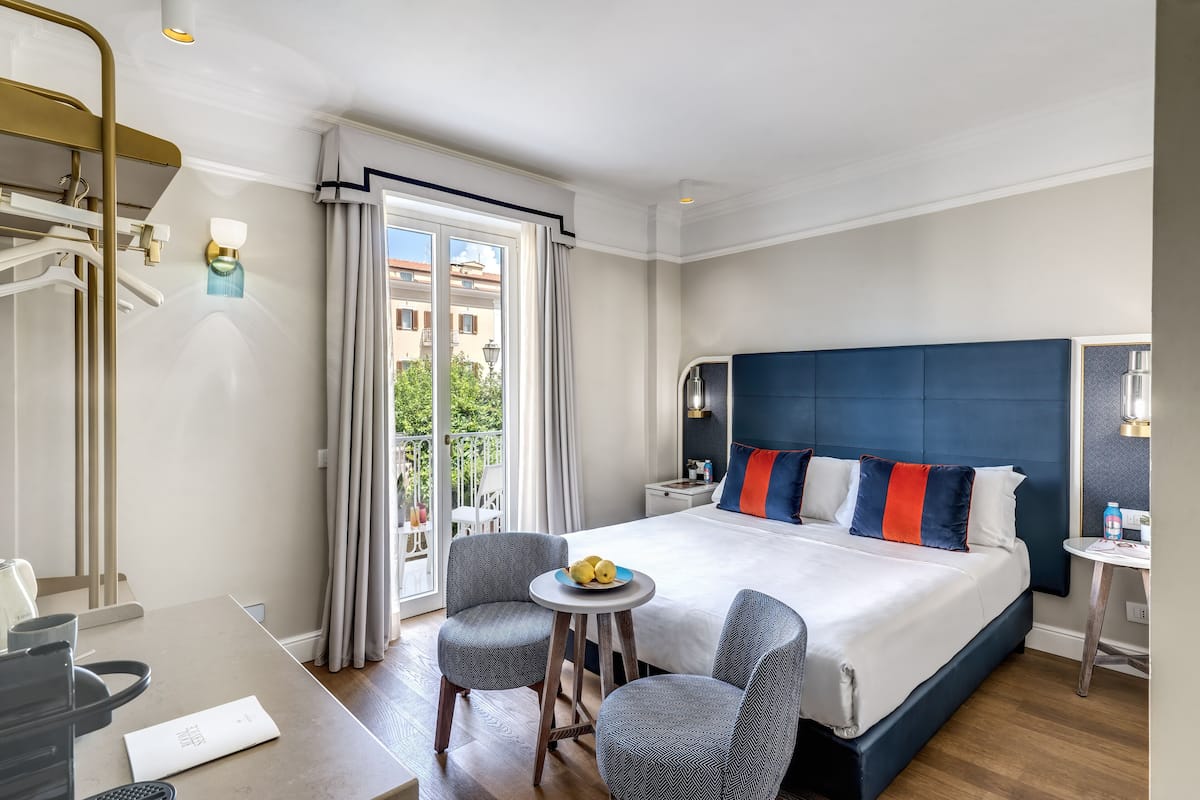 Ett modernt hotellrum med en stor säng, ett litet bord, två stolar, ett skrivbord och en balkong med gardiner.