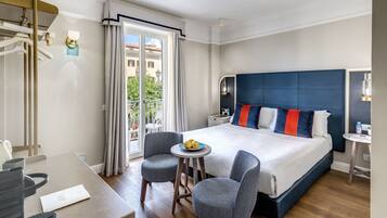 Quarto de hotel moderno com uma cama grande, uma mesa pequena, duas cadeiras, uma escrivaninha e uma varanda com cortinas.