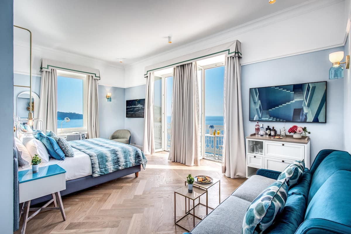 Ett modernt hotellrum med en säng, en soffa, ett skrivbord och en TV. Rummet har stora fönster med gardiner och utsikt över havet.
