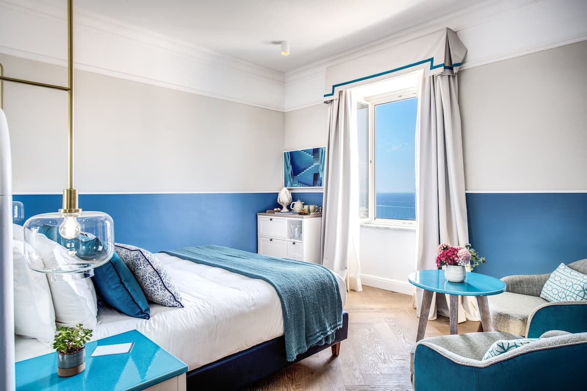 Ett hotellrum med en säng, ett litet bord med en vas med blommor, en stol och utsikt över havet.