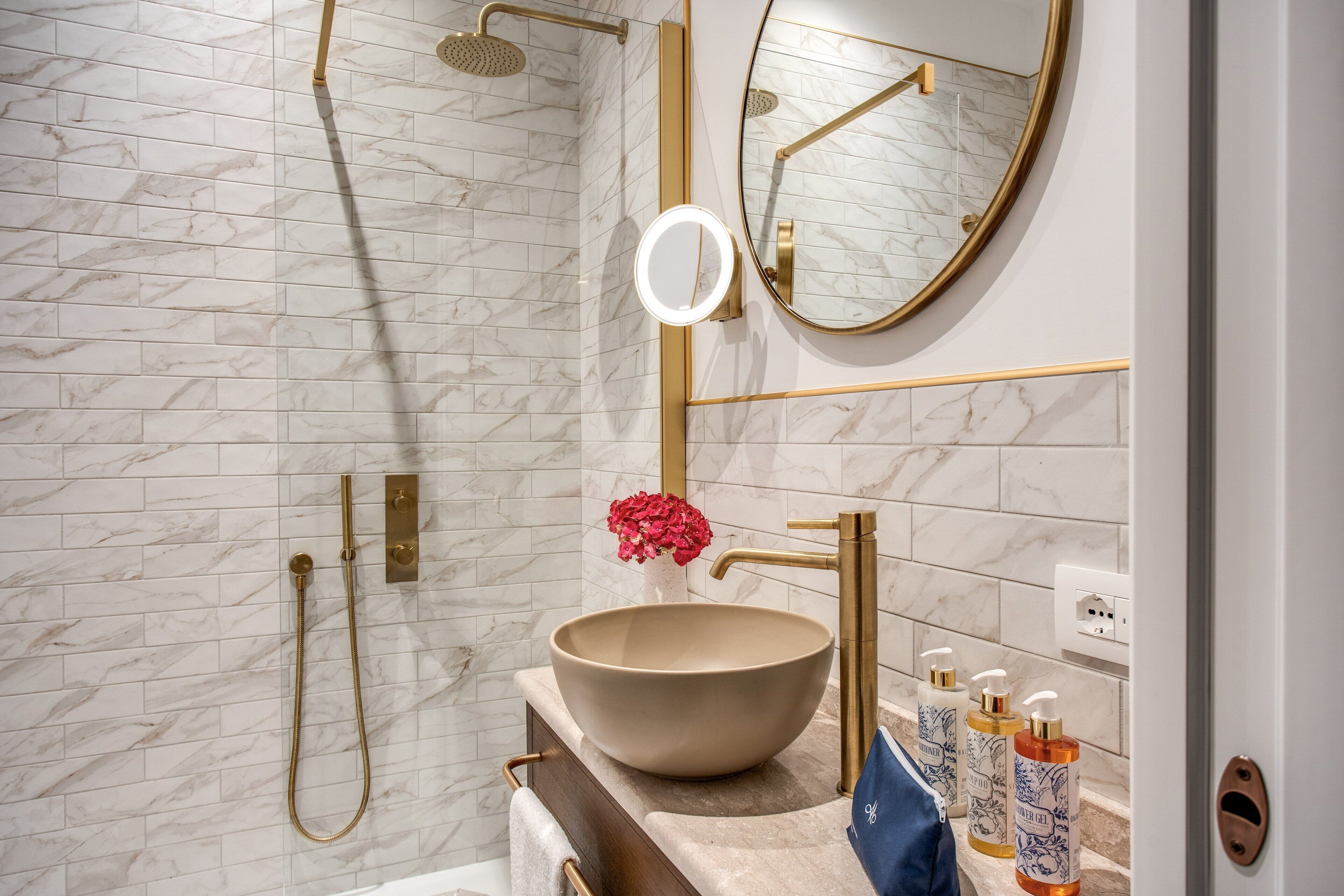 A modern bathroom with a marble wall, a large round mirror, a beige sink, and gold fixtures.
