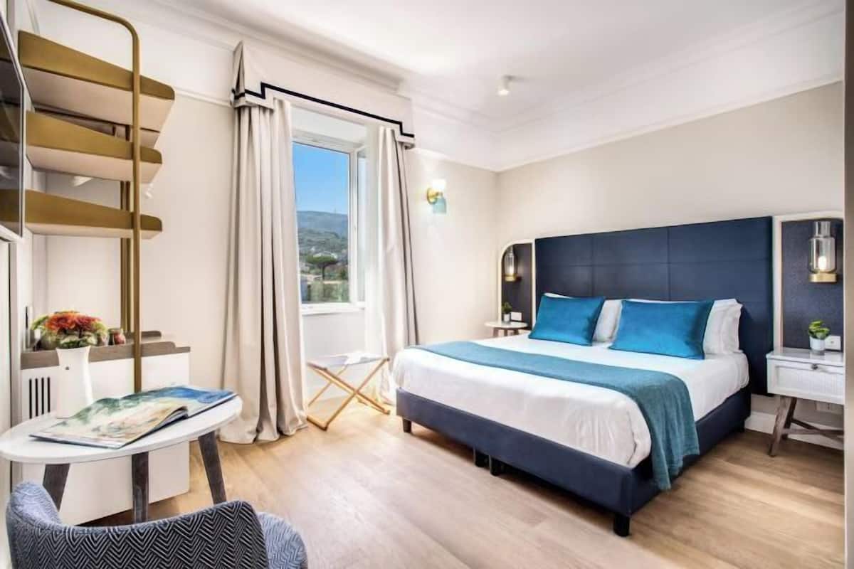 Ett hotellrum med en säng, ett skrivbord, en stol, ett fönster med gardiner och utsikt över omgivningen.