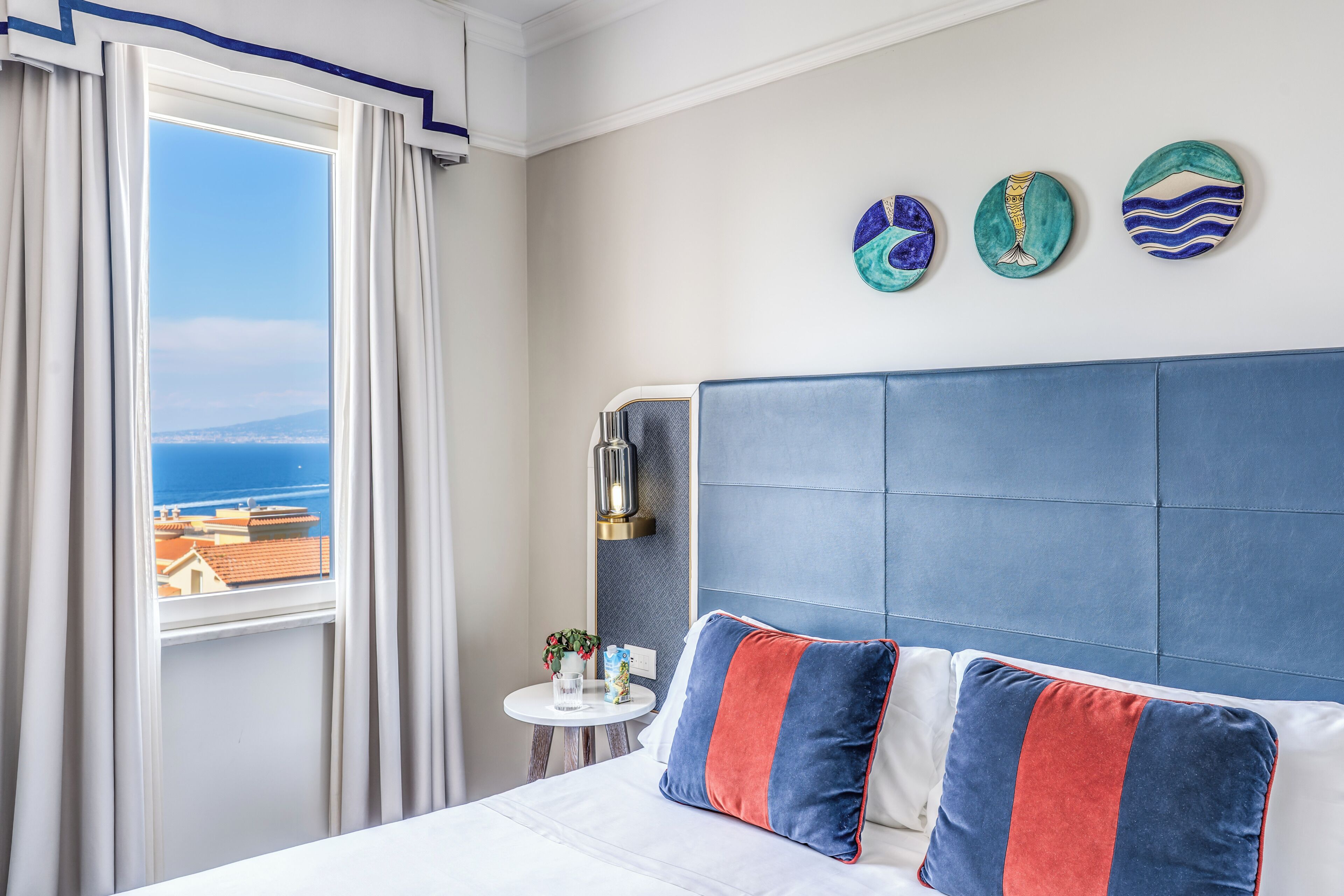 A hotel room with a bed, a view of the sea, a small table with a plant, and decorative plates on the wall.