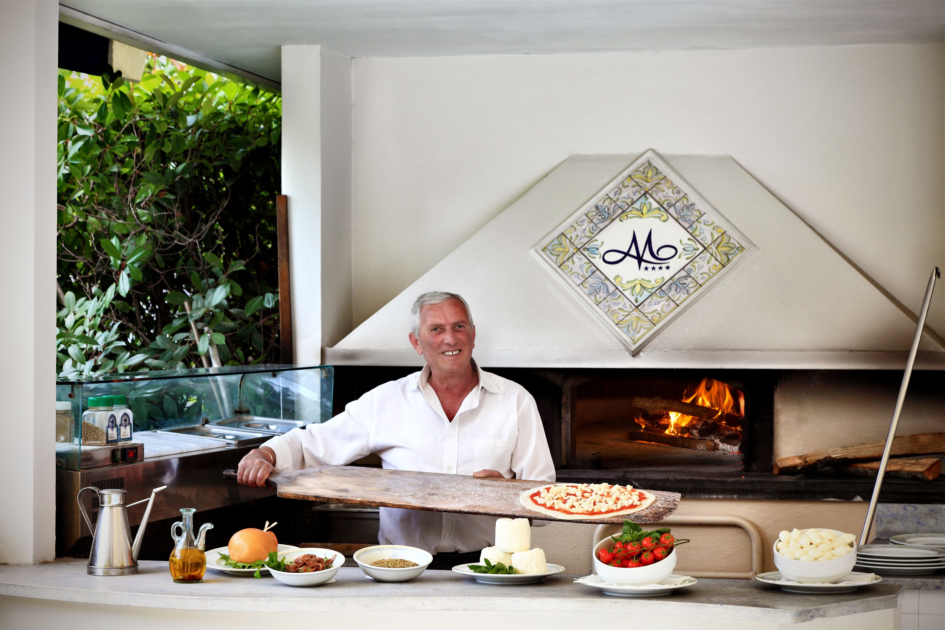 A chef holding a pizza in front of a wood-fired oven with a decorative wall sign.
