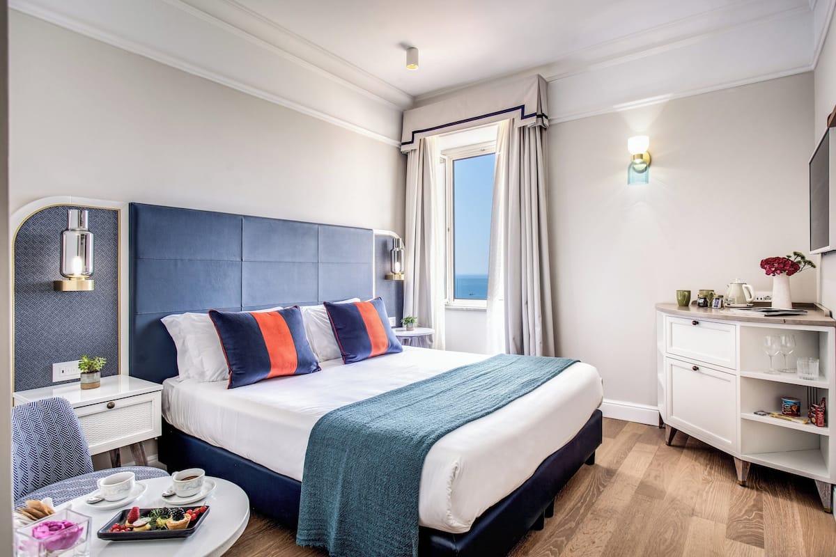 Ett hotellrum med en stor säng, ett nattduksbord med en lampa, ett litet bord med ett bricka med mat och utsikt över havet genom ett fönster.