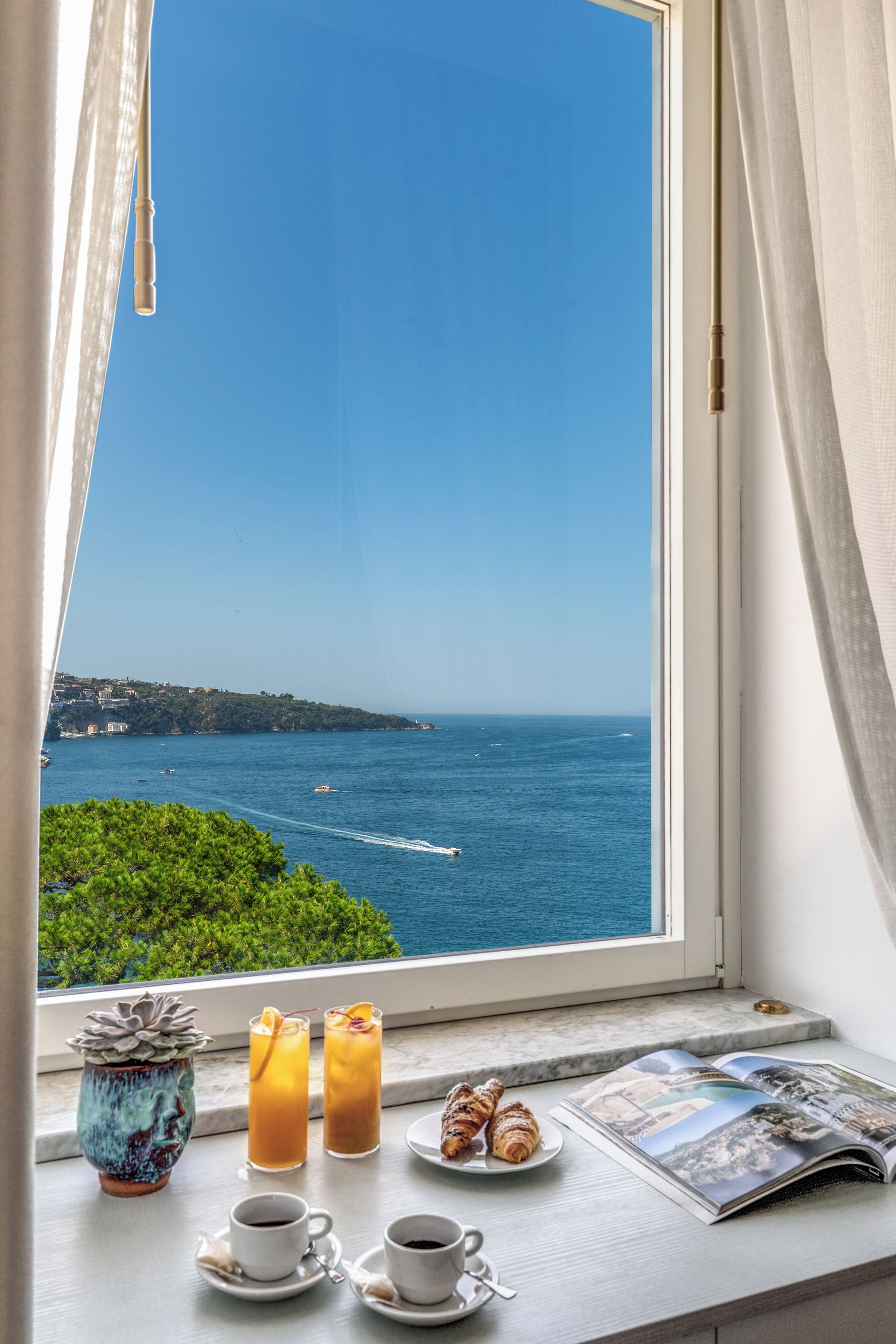 Una ventana con vista al mar, una mesa con bebidas, un croissant y una planta exuberante.