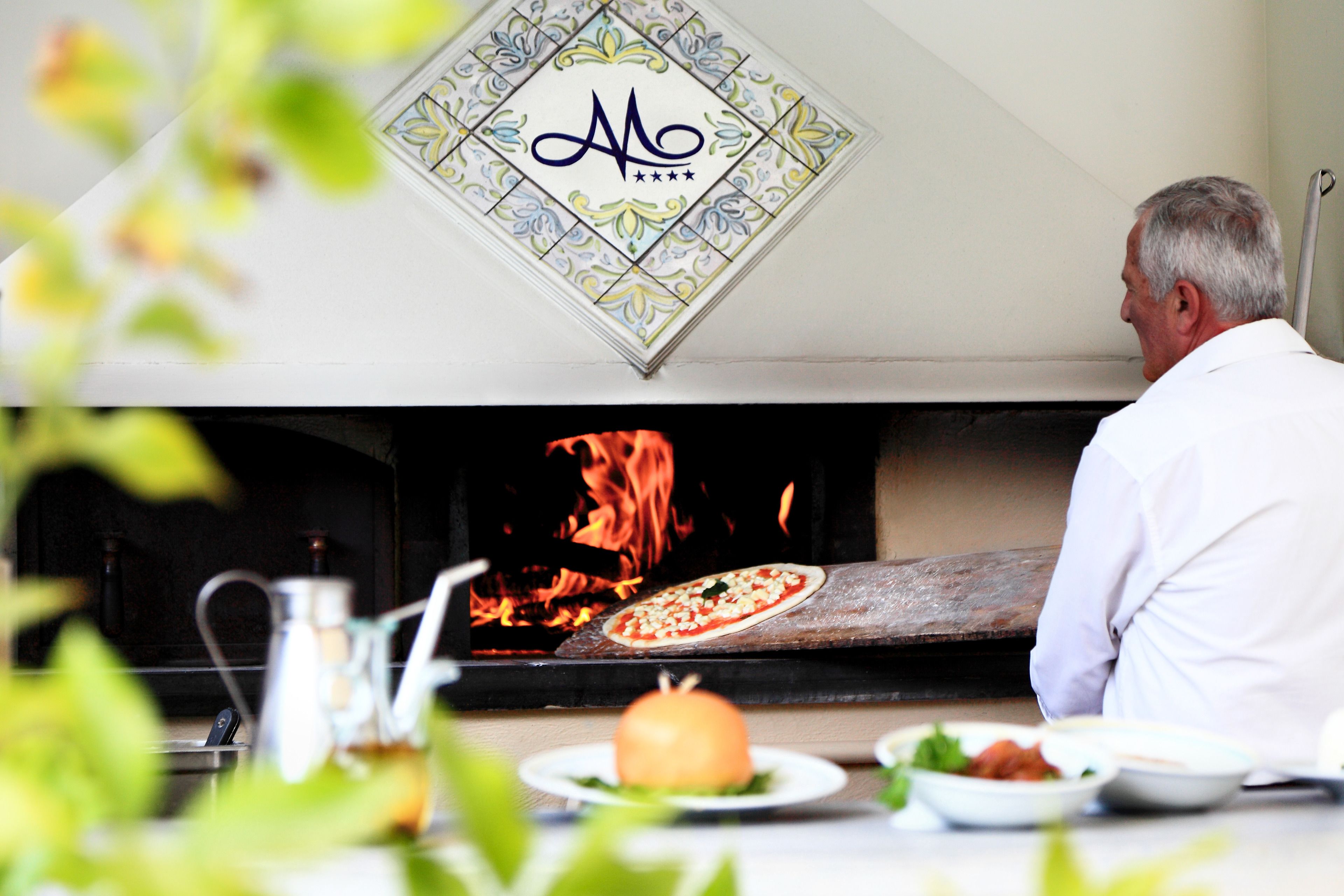 Un uomo con una camicia bianca osserva una pizza che cuoce in un forno a legna.