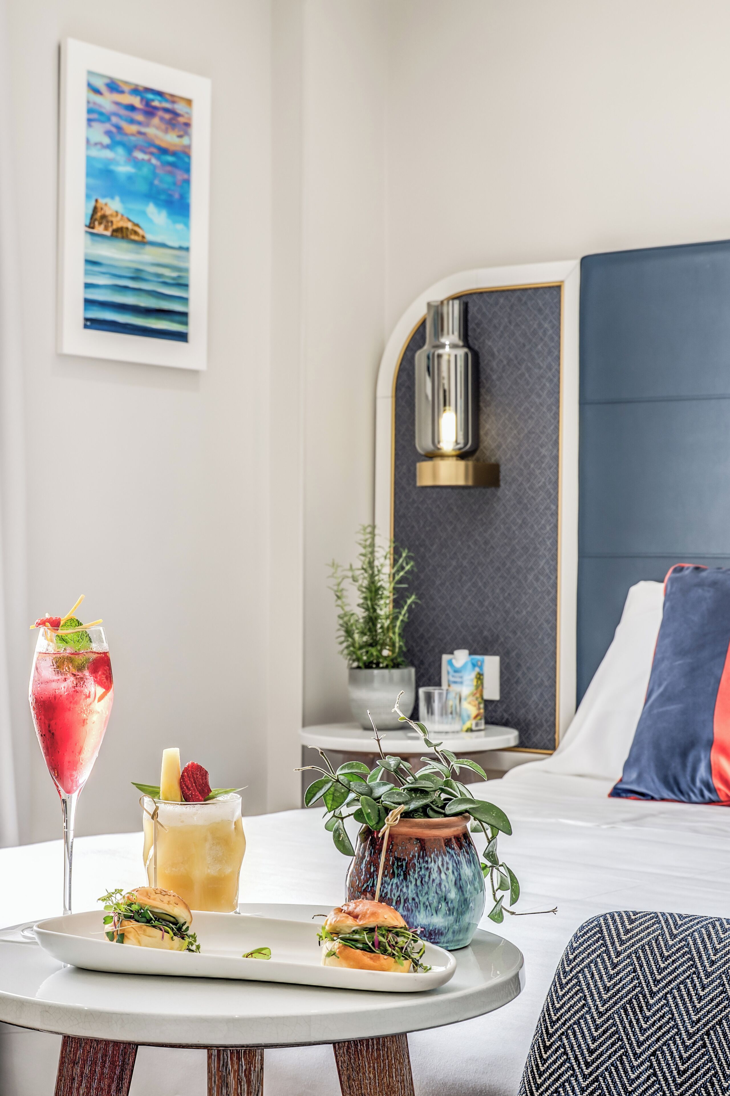 A hotel room with a bed, a bedside table with a plant, a glass of drink, and a plate with food.