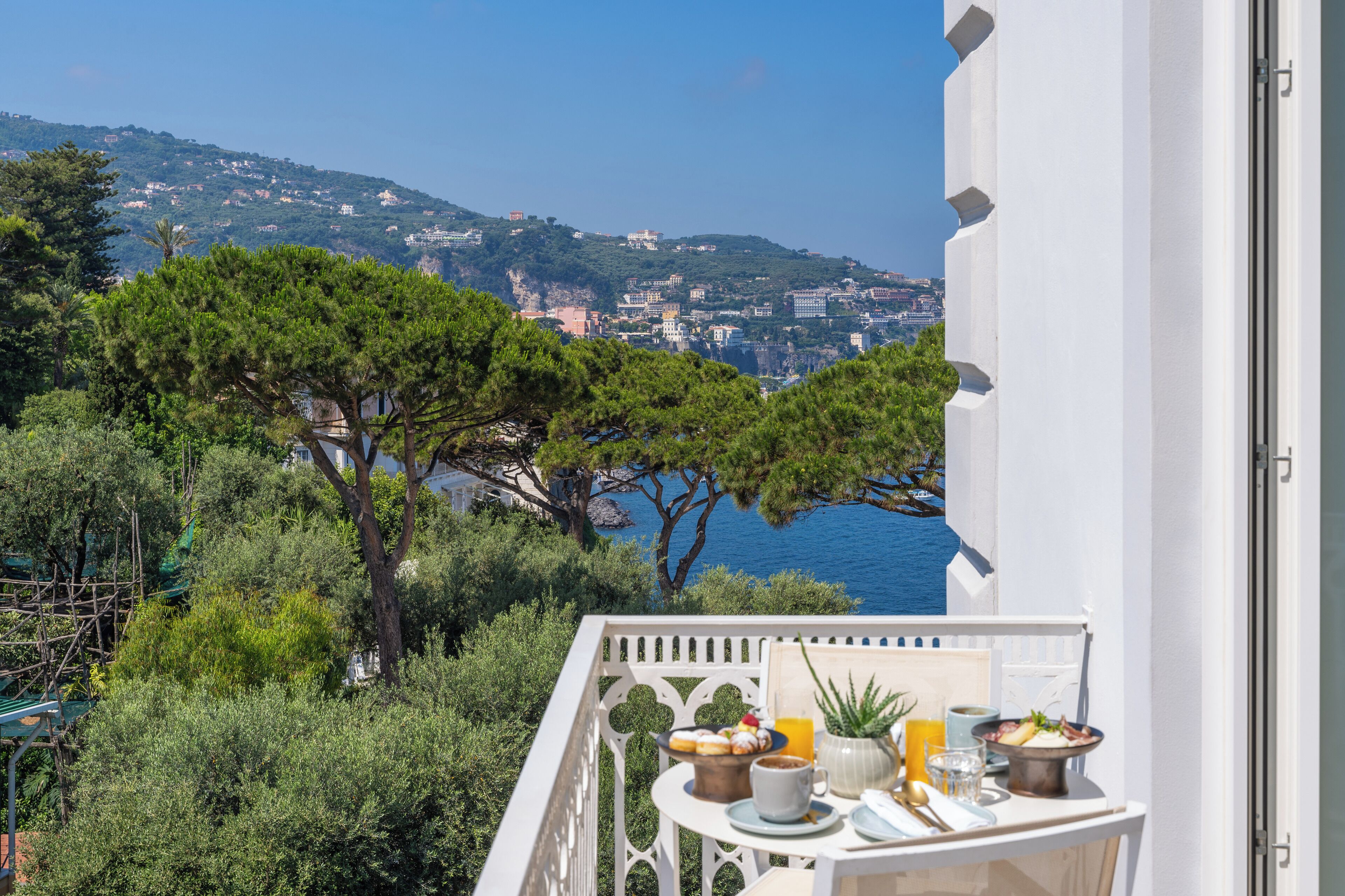 Un balcone con tavolo apparecchiato per la colazione, vista su una cittadina costiera e il mare.
