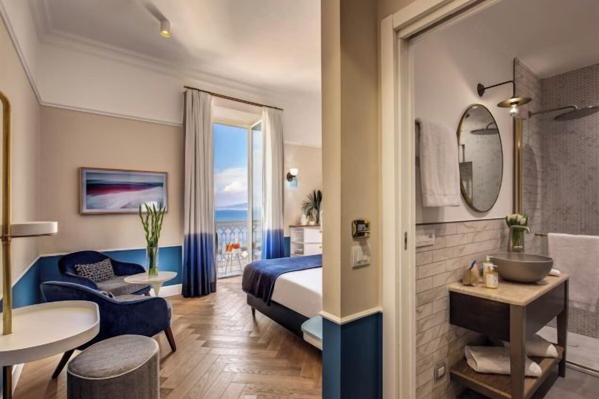 A hotel room with a bed, a seating area with a sofa and chairs, a small table, a bathroom with a sink and mirror, and a view through a door.