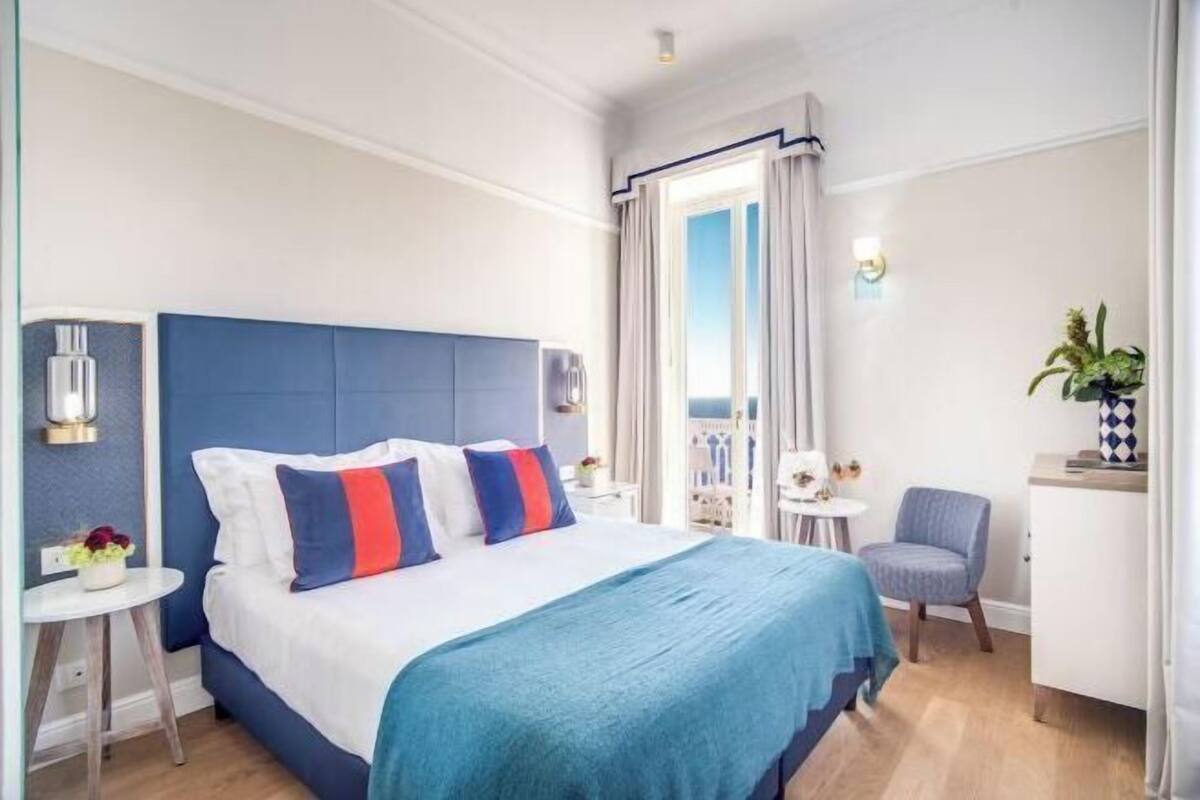 Ett hotellrum med en säng, ett skrivbord, en stol, ett litet bord med en växt och utsikt över omgivningen.