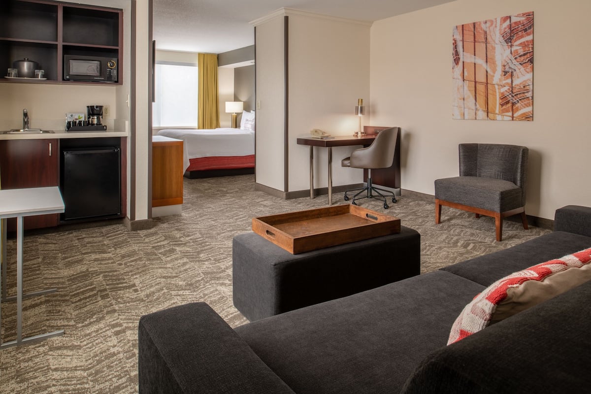 A hotel room with a kitchenette, a bed, a desk, two chairs, and a large painting on the wall.