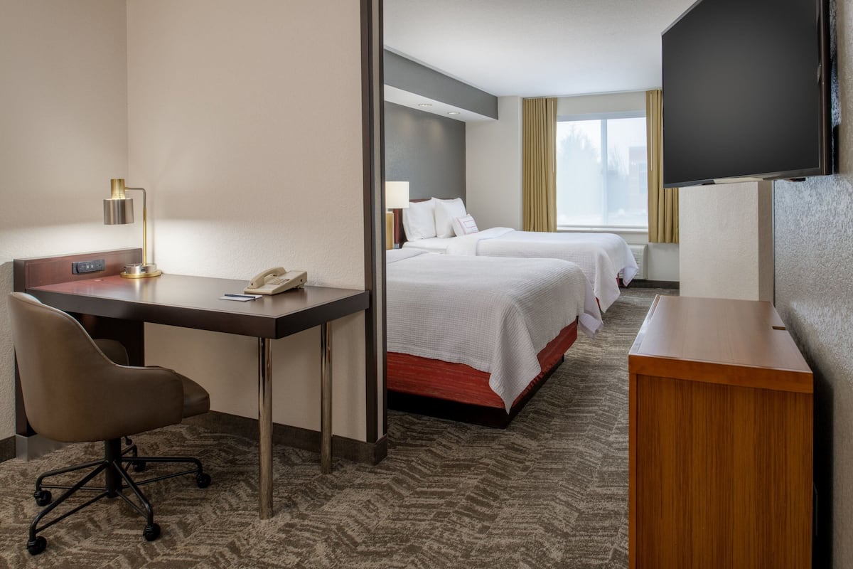 A hotel room with a desk, chair, two beds, a TV, and a window with curtains.