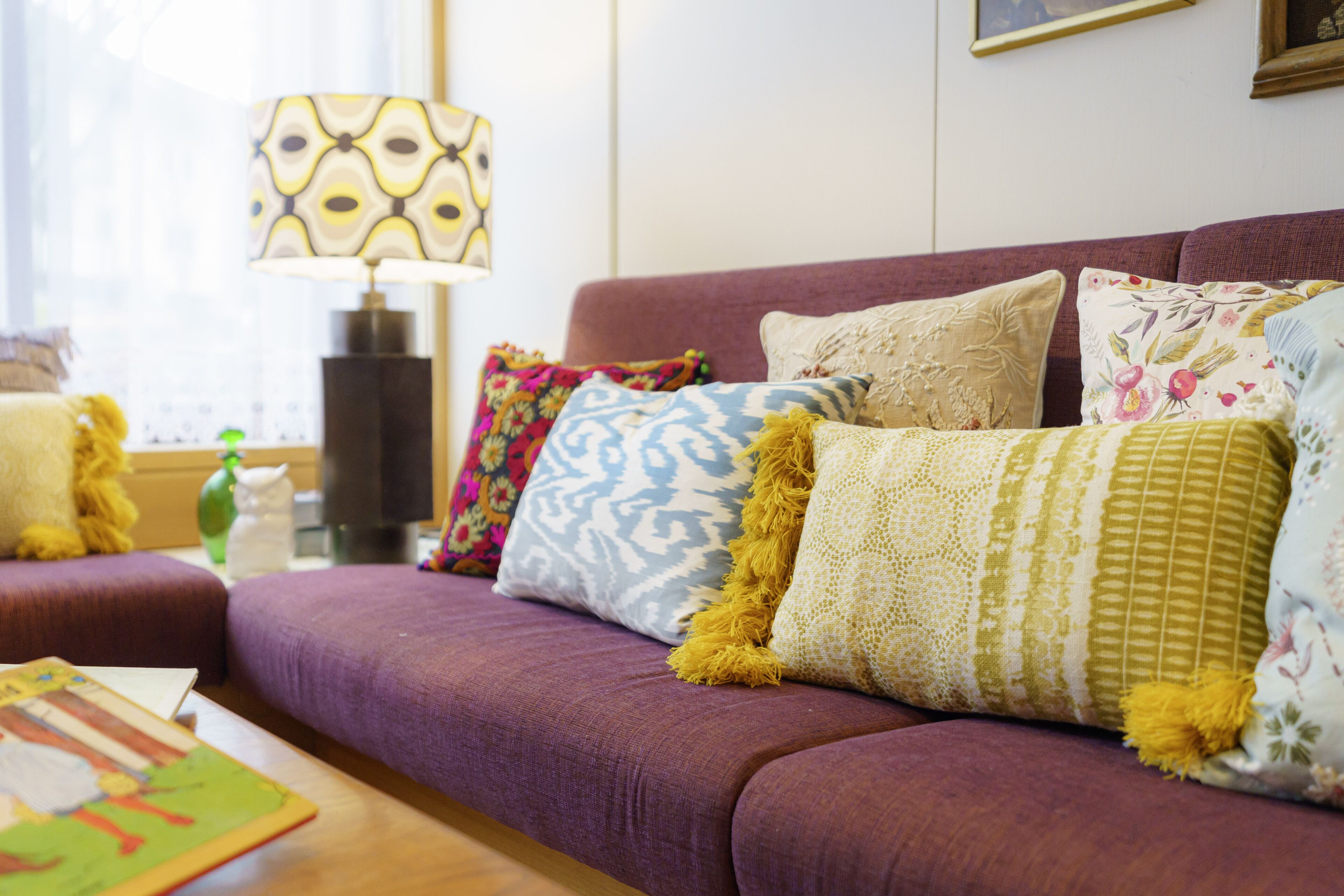 A purple sofa with patterned cushions, a lamp, and a book on a wooden table.