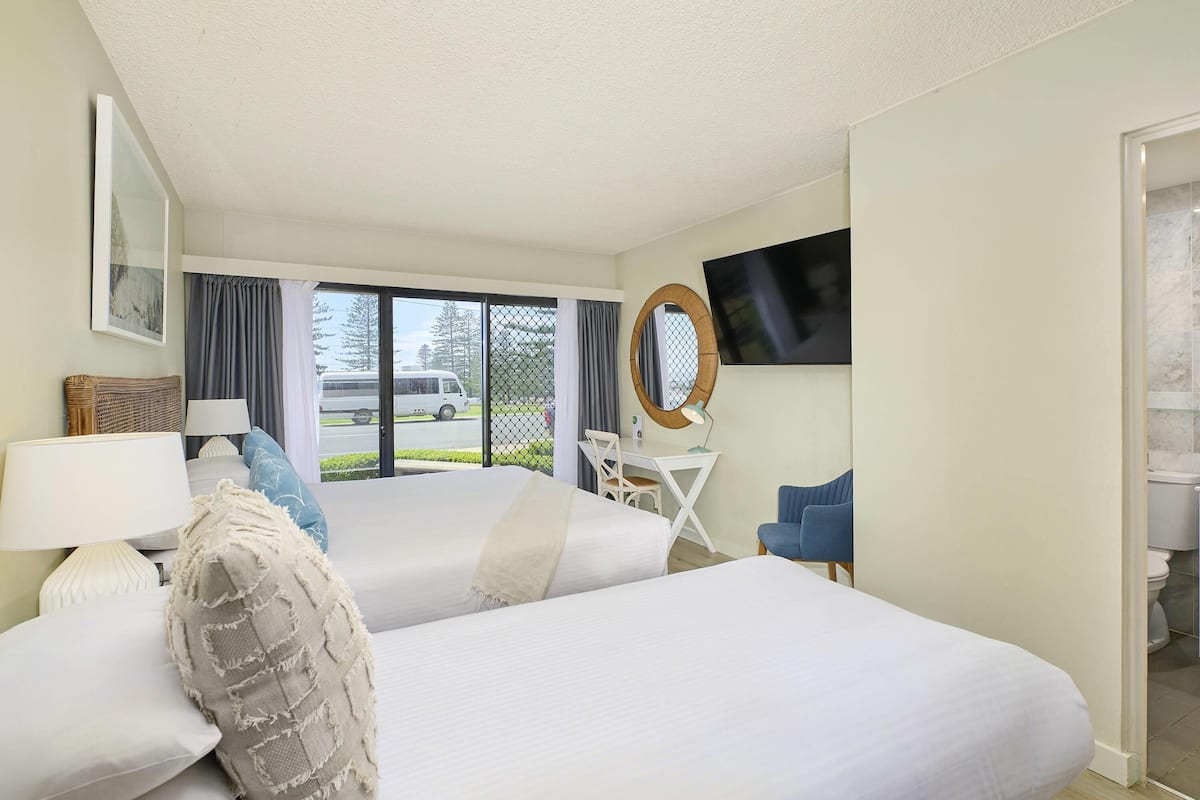 A hotel room with two beds, a desk with a chair, a TV, and a view of the street outside.