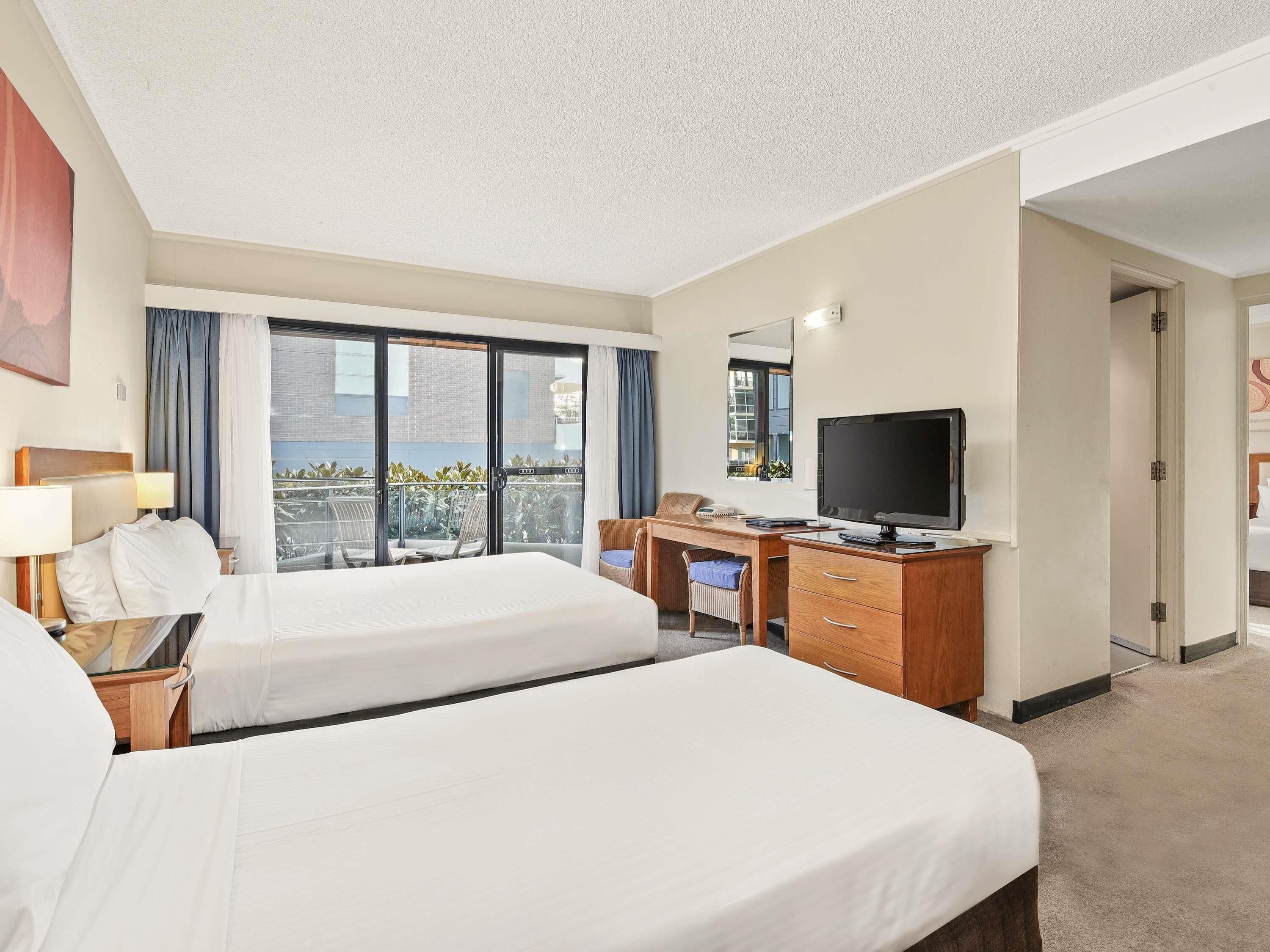A hotel room with two beds, a desk with a TV, a chair, and a balcony with curtains.