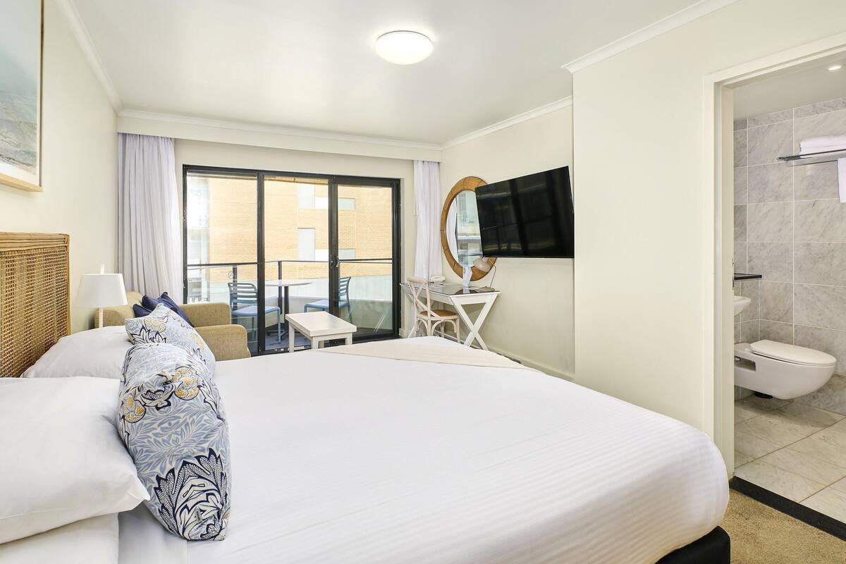 A hotel room with a large bed, a desk with a chair, a TV mounted on the wall, and a bathroom with a toilet and sink.