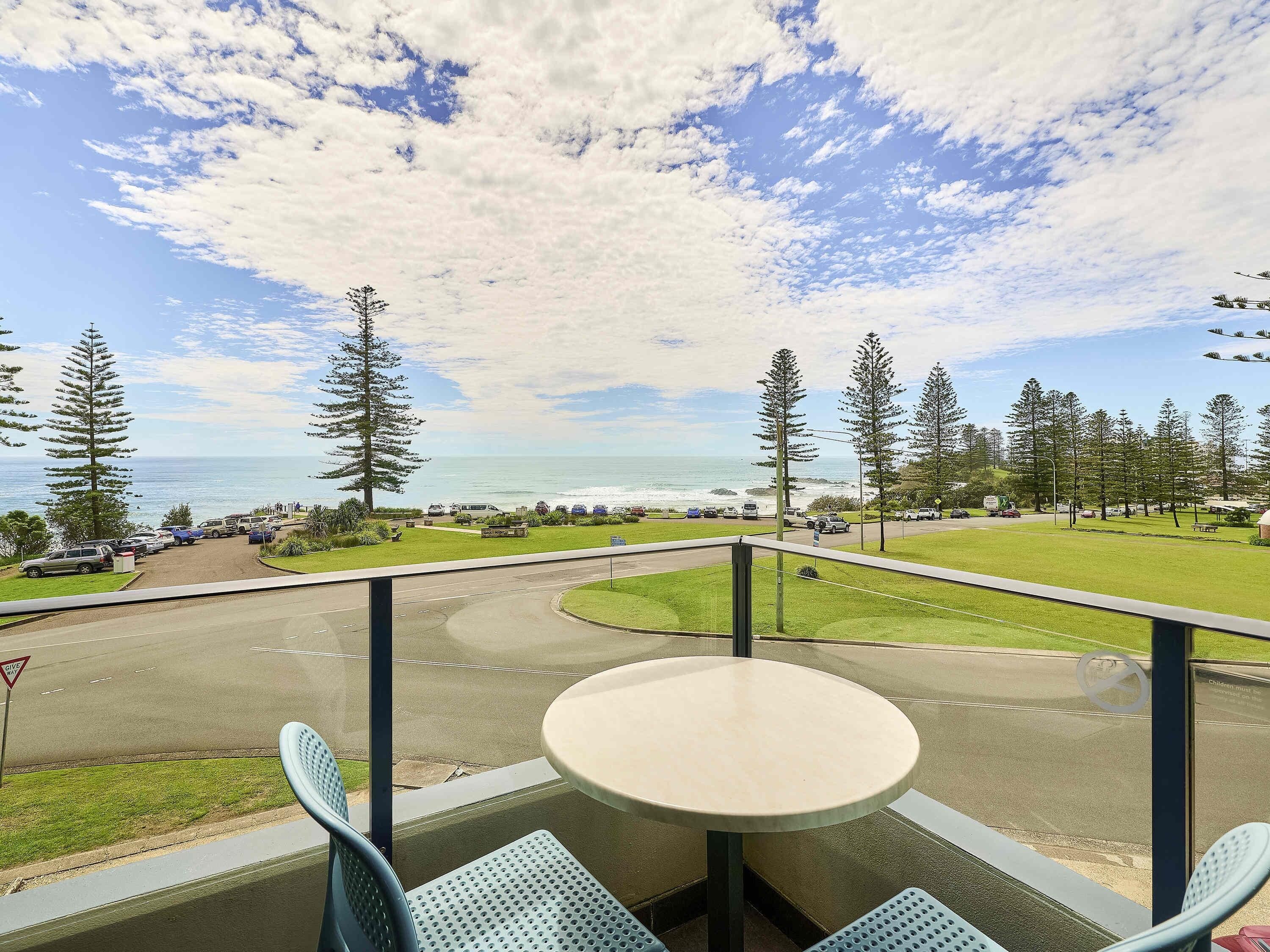 A balcony with a table and chairs overlooking a road with parked cars, a grassy area, and a view of the ocean.