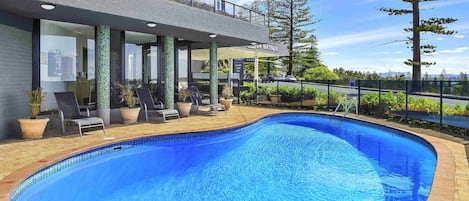 A swimming pool with lounge chairs, a covered area with a sign, and a view of trees and a road.