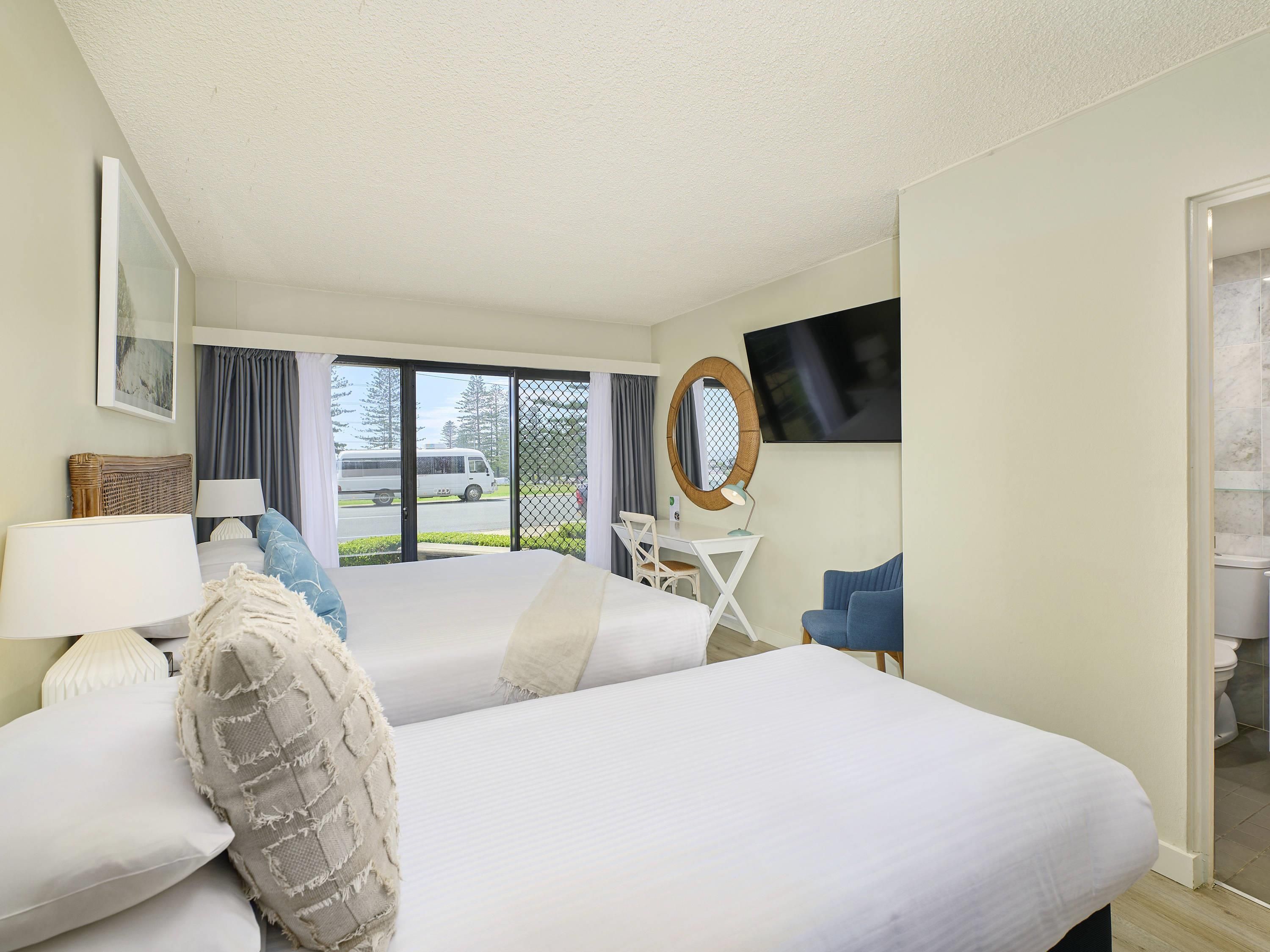 Habitación de hotel con dos camas, un escritorio con silla, televisión y vista a la calle.