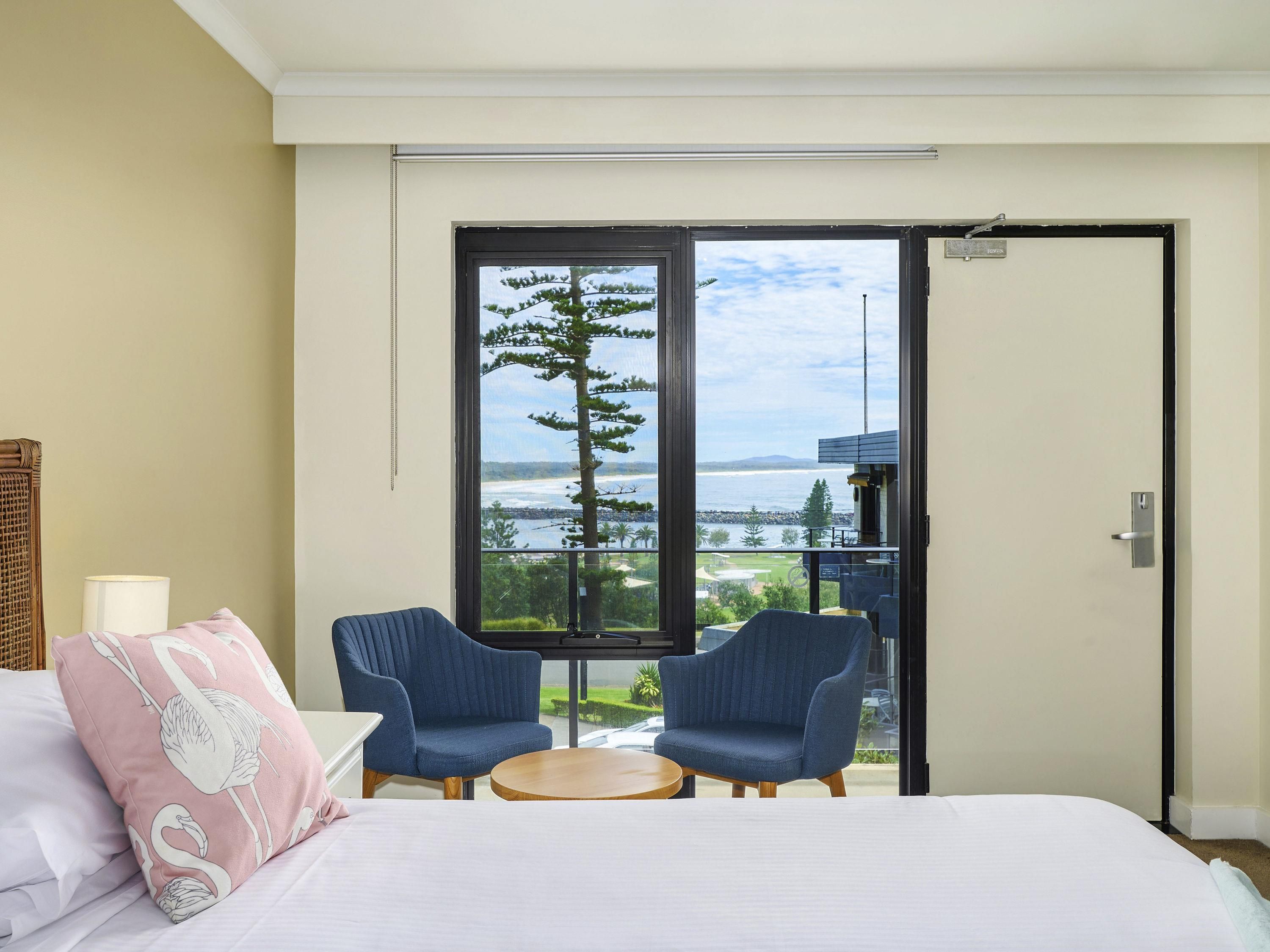 A bedroom with a large bed, two chairs, a small table, and a window with a view of the sea and trees.