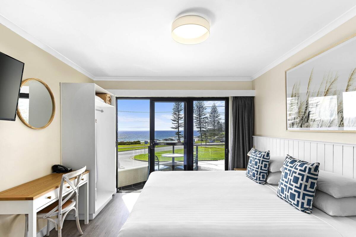A hotel room with a bed, a desk, a chair, a television, and a large window with a view of the sea and trees.