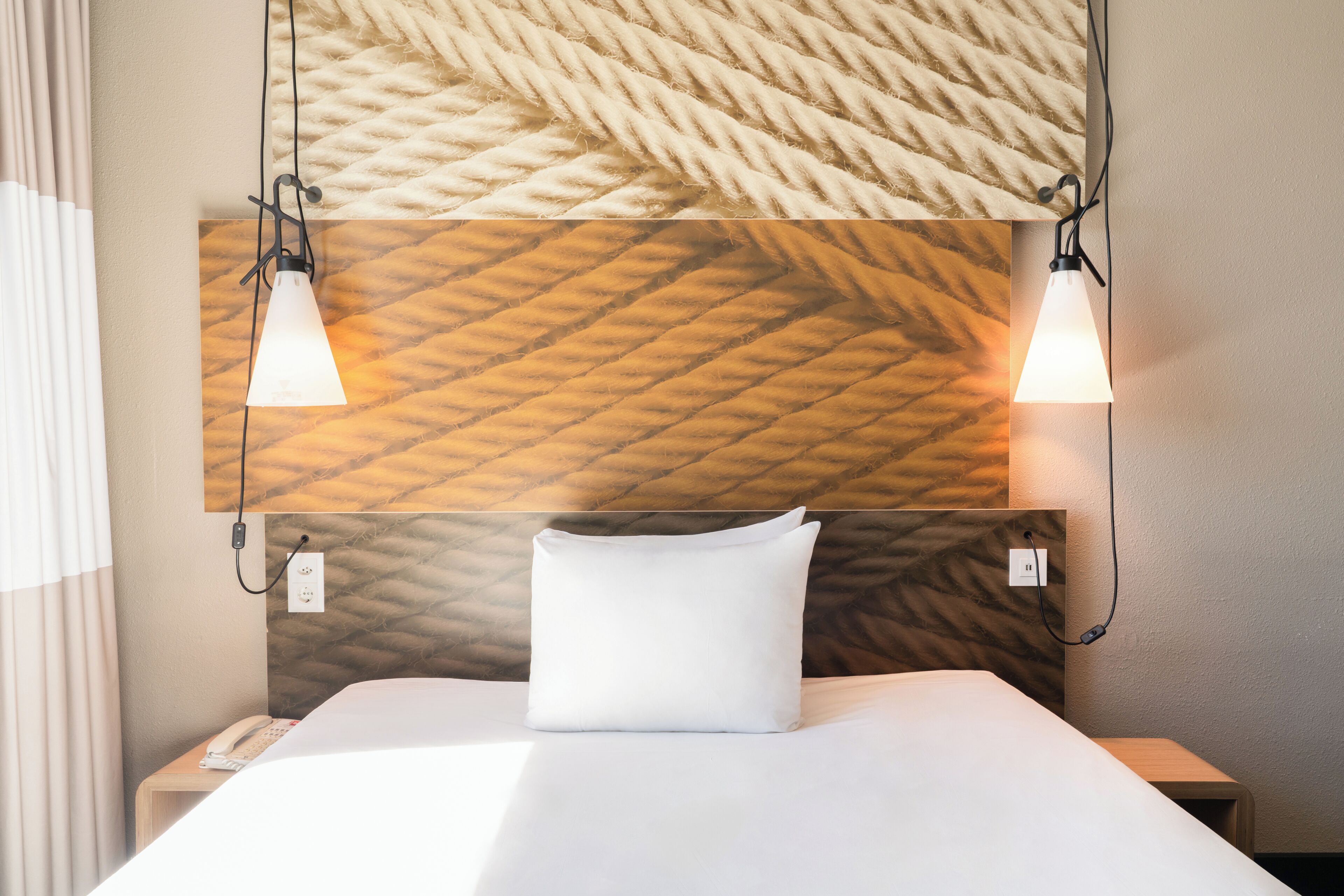 Habitación de hotel con cama, teléfono en la mesita de noche, cabecero con textura y dos lámparas de pared.