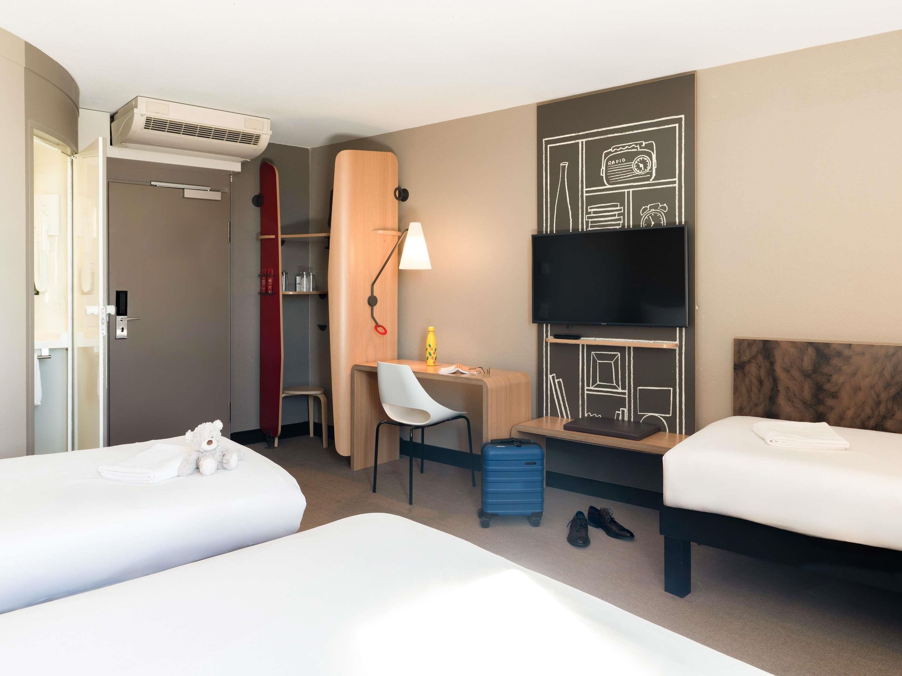 A hotel room with two beds, a desk with a chair, a TV, a wardrobe, and a suitcase.
