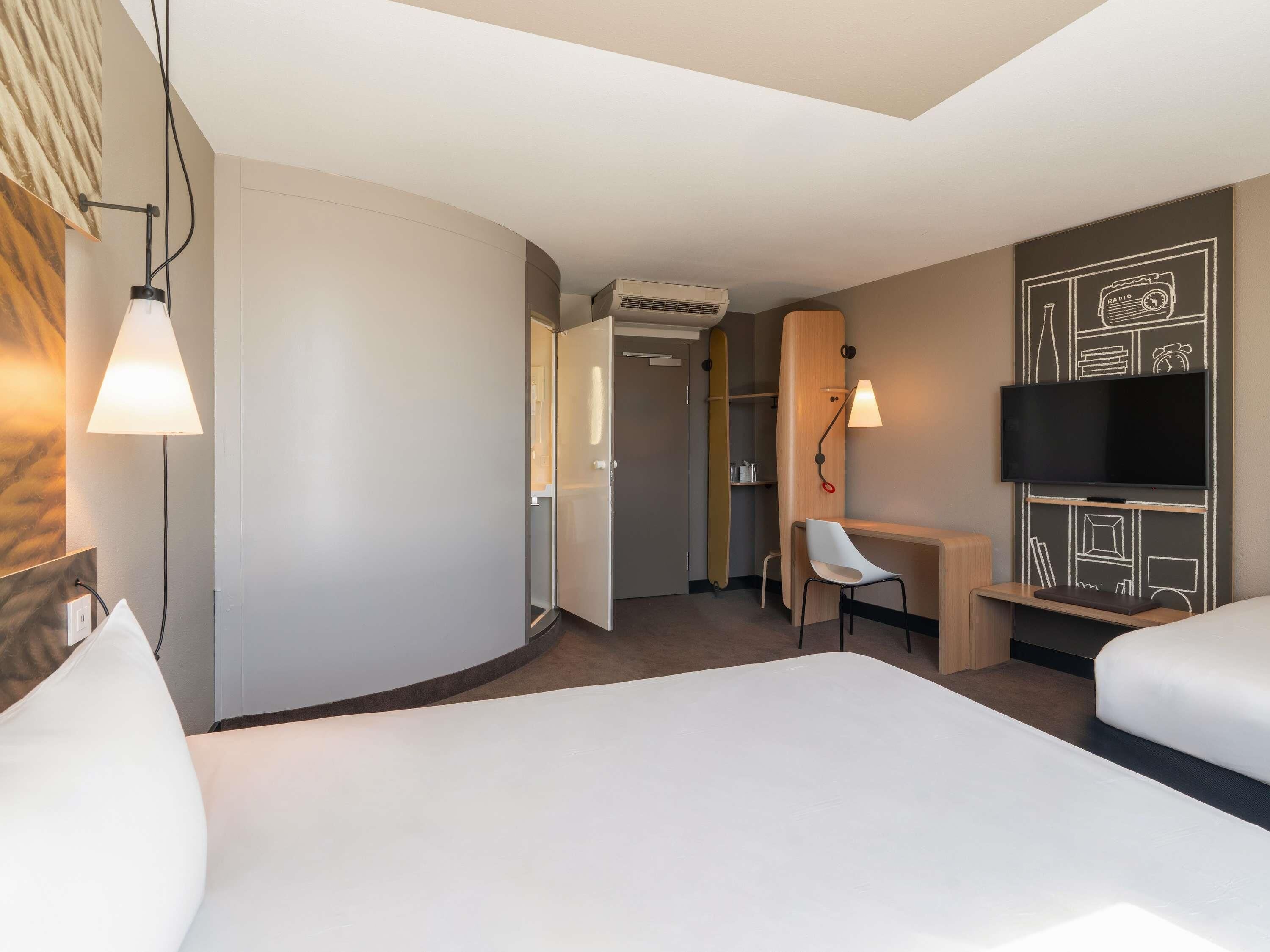 A hotel room with a bed, a desk, a chair, a TV, and an air conditioning unit.